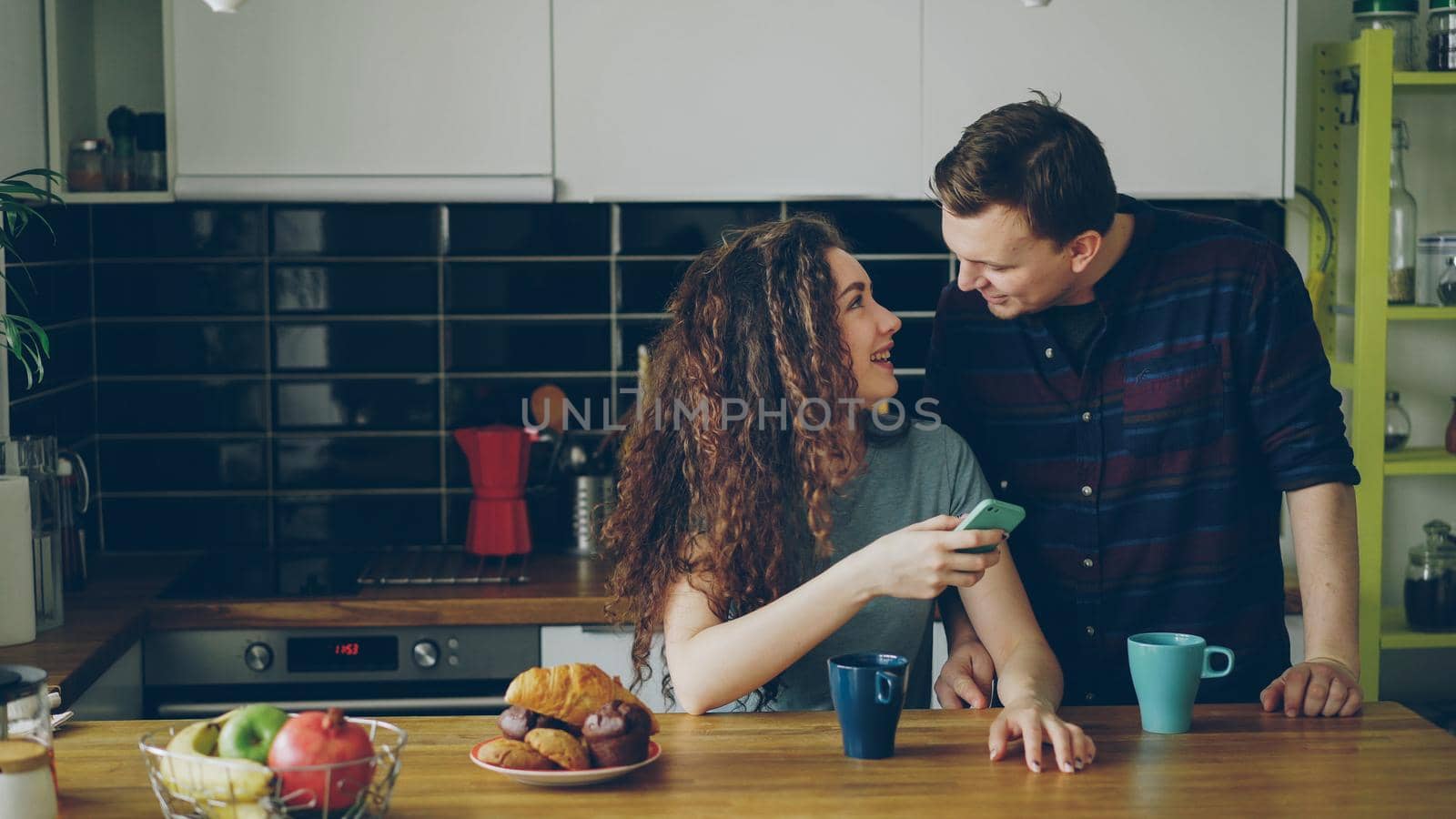 young attractive curly happy caucasian woman is sitting in kitchen texting someone via smartphone, her husband comes and she shows something funny in phone by silverkblack