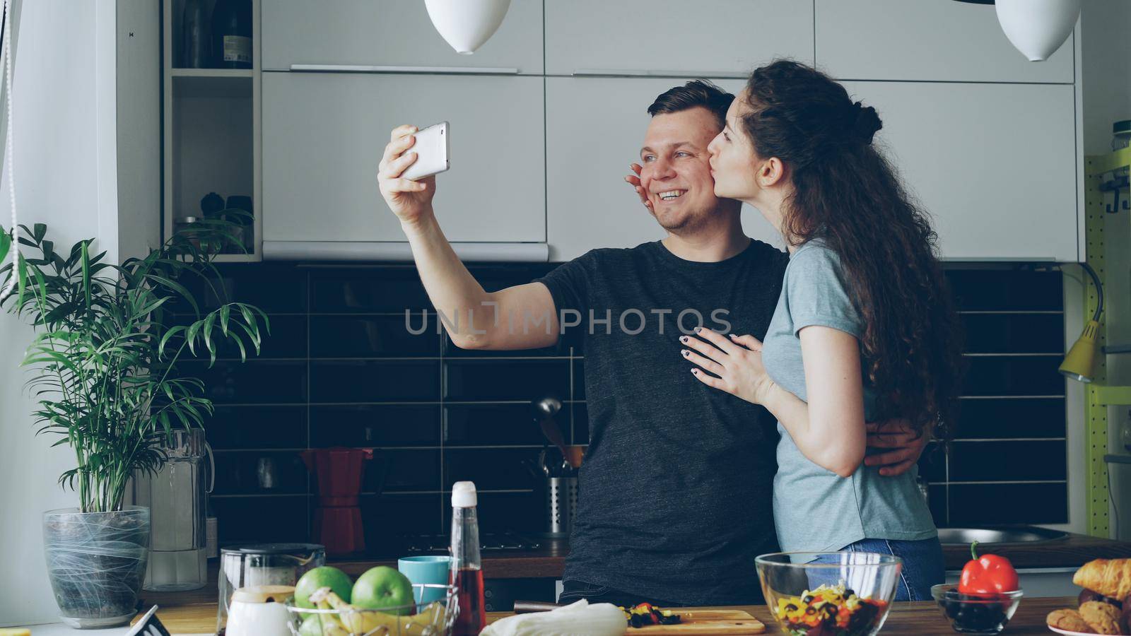 Young funny couple taking self portrait with smartphone camera while cooking in the kitchen at home in the morning
