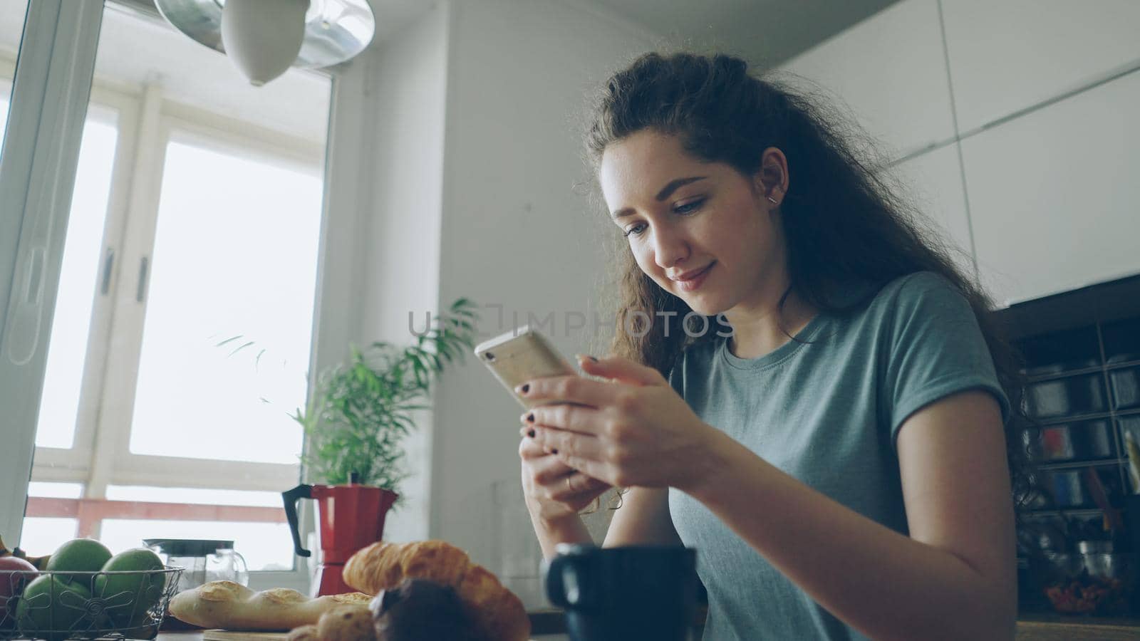 young beautiful curly pretty caucasian woman sitting at table in nice kitchen using smartphone, she is texting someone and smiling, calm and happy by silverkblack