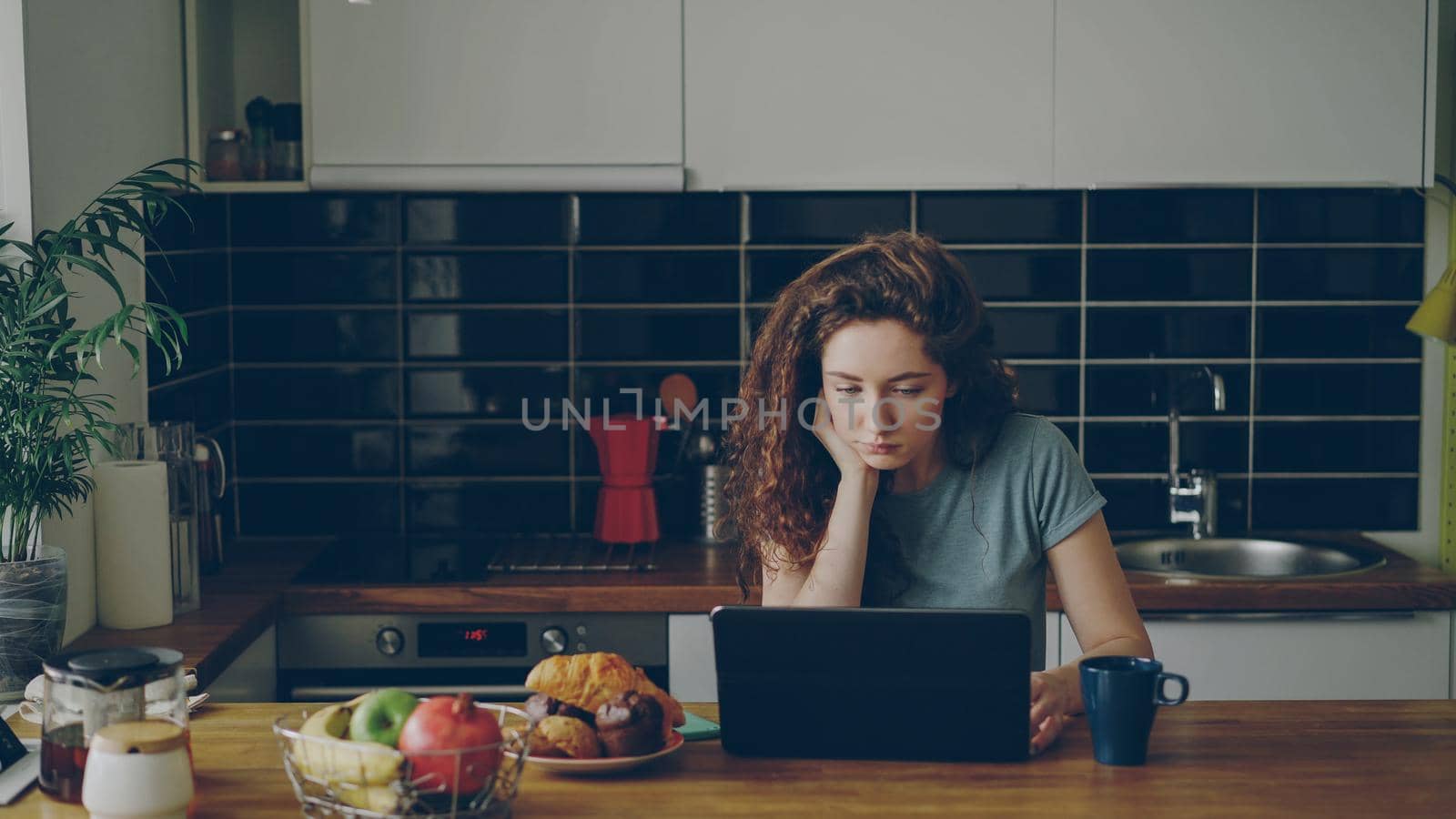 Young thoughtful curly caucasian woman is sitting in modern kitchen indoors in front of laptop, she takes a glance into window, she is thinking somenthing over