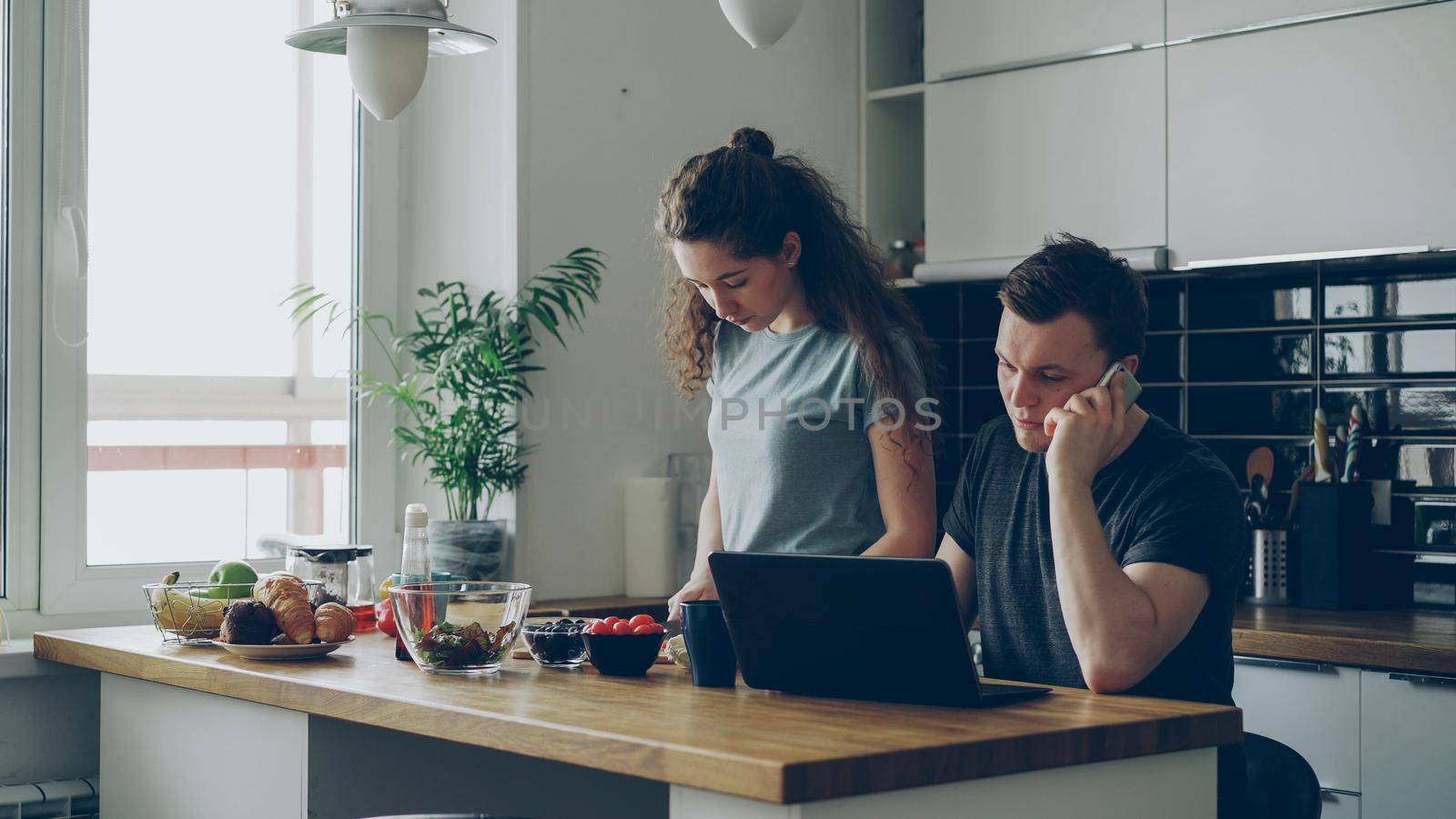 serious couple in kitchen, woman is cutting salad, she is standing silent, man is sitting at table working on laptop and talking on phone, he is annoyed and angry