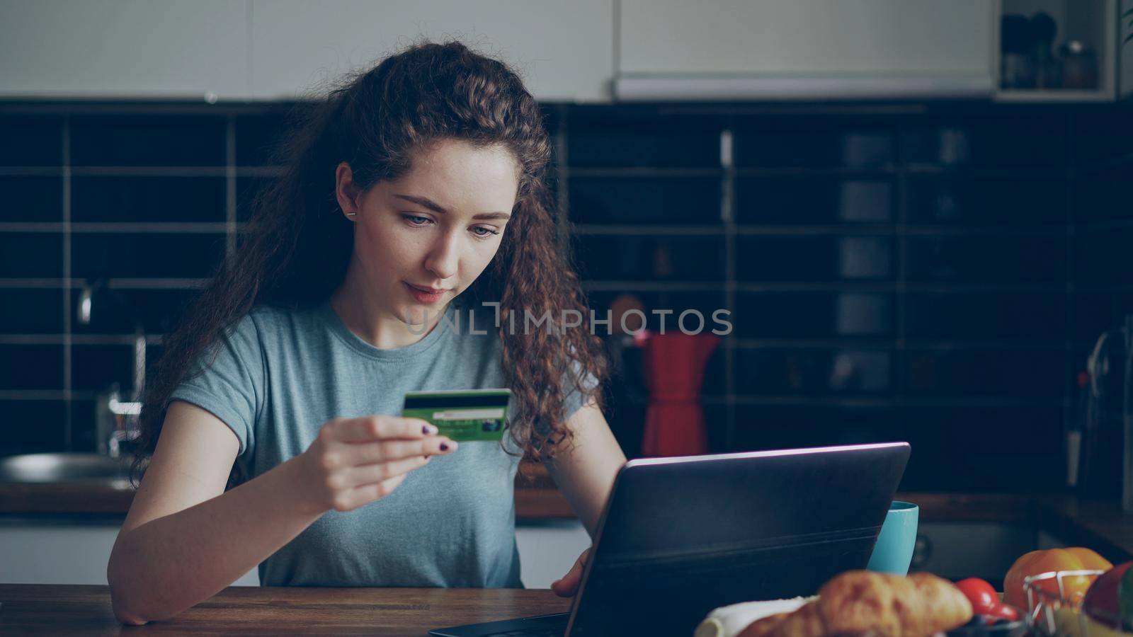 Cheerful young woman having online shopping using laptop computer and credit card while have breakfast in the kitchen at home by silverkblack