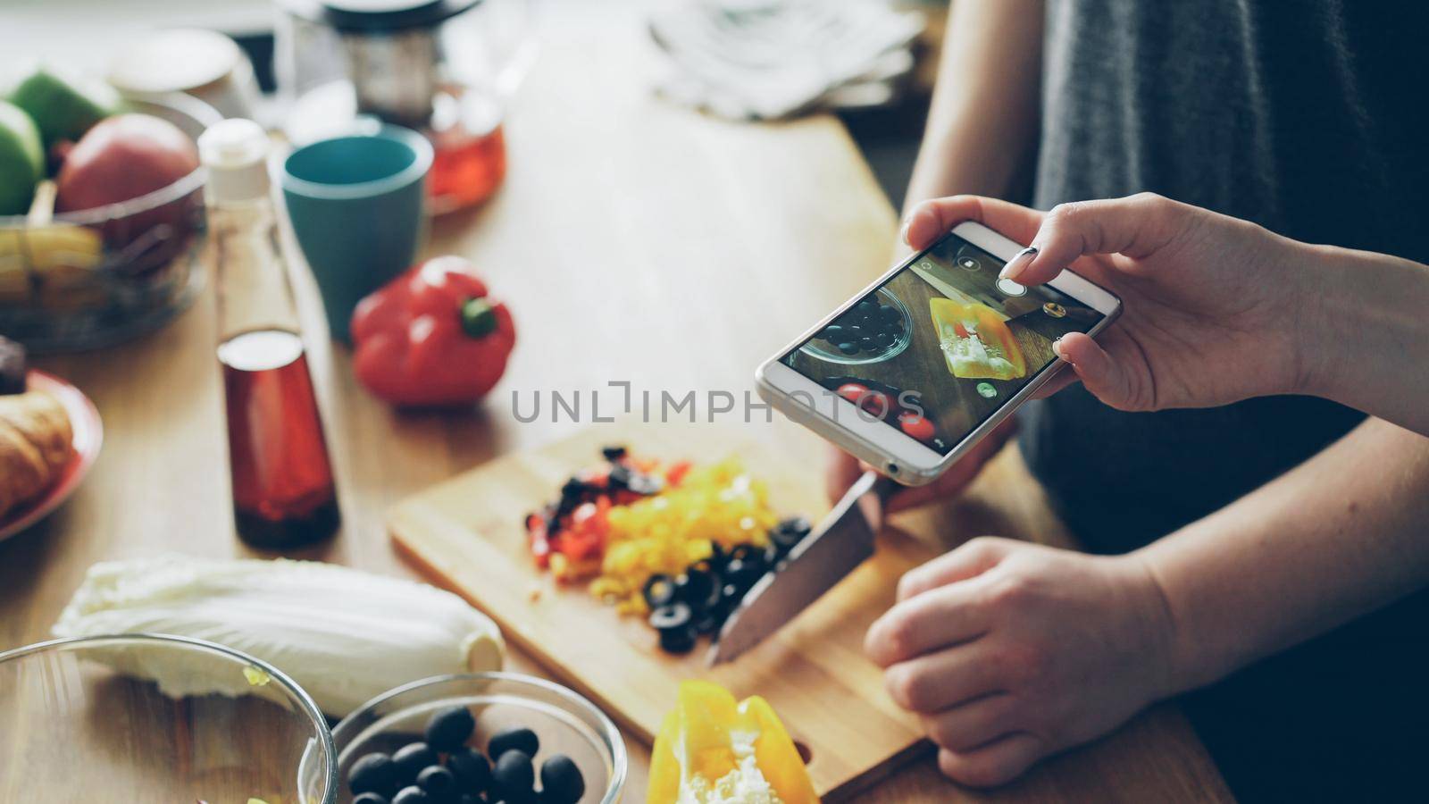 Close-up of man hands cutting vegetables for salad and his girlfriend taking photos on smartphone camera for social media in the kitchen at home by silverkblack
