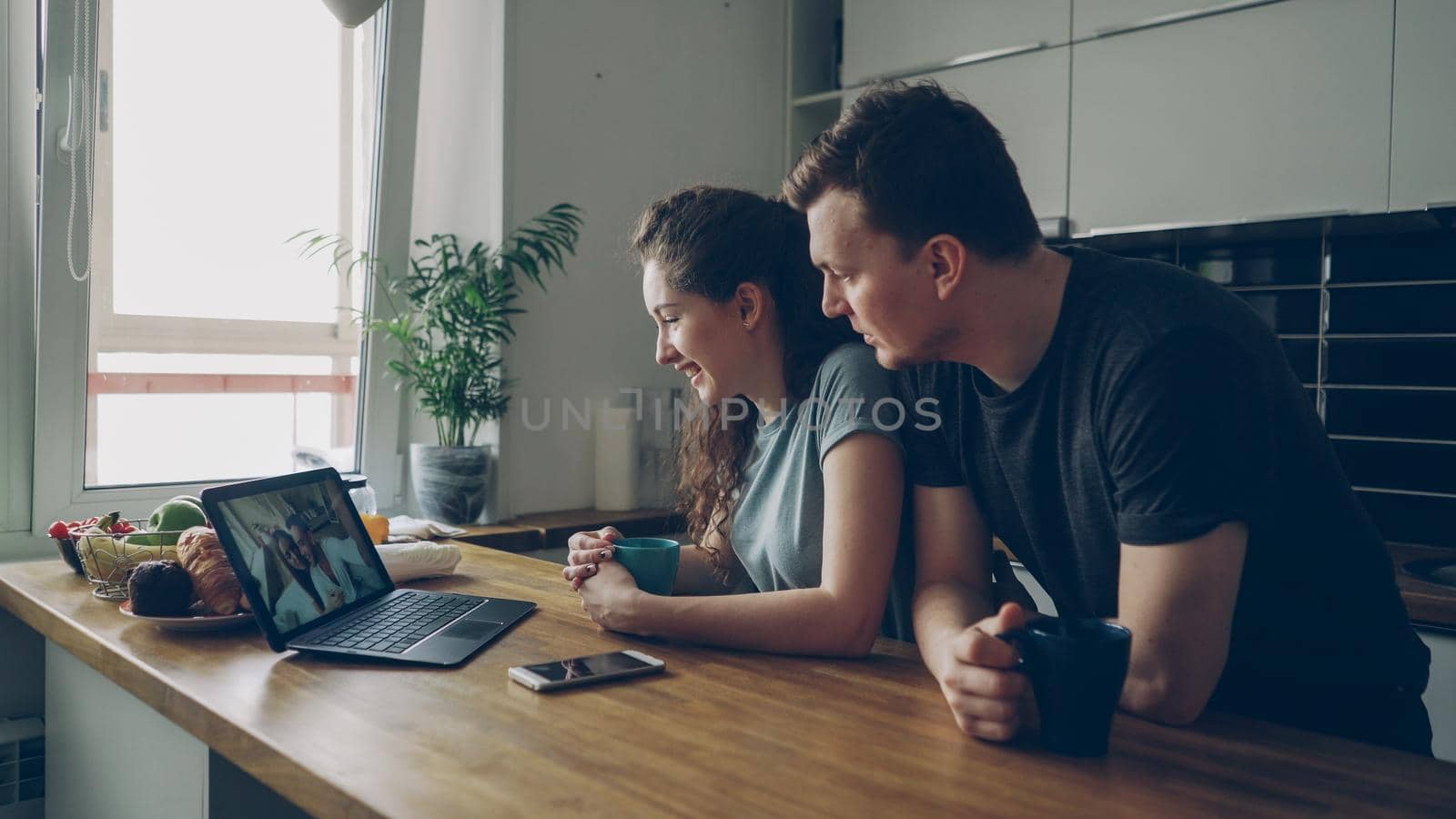 Young caucasian couple sitting at table in front of laptop talking with their happy positive tanned friends from abroad