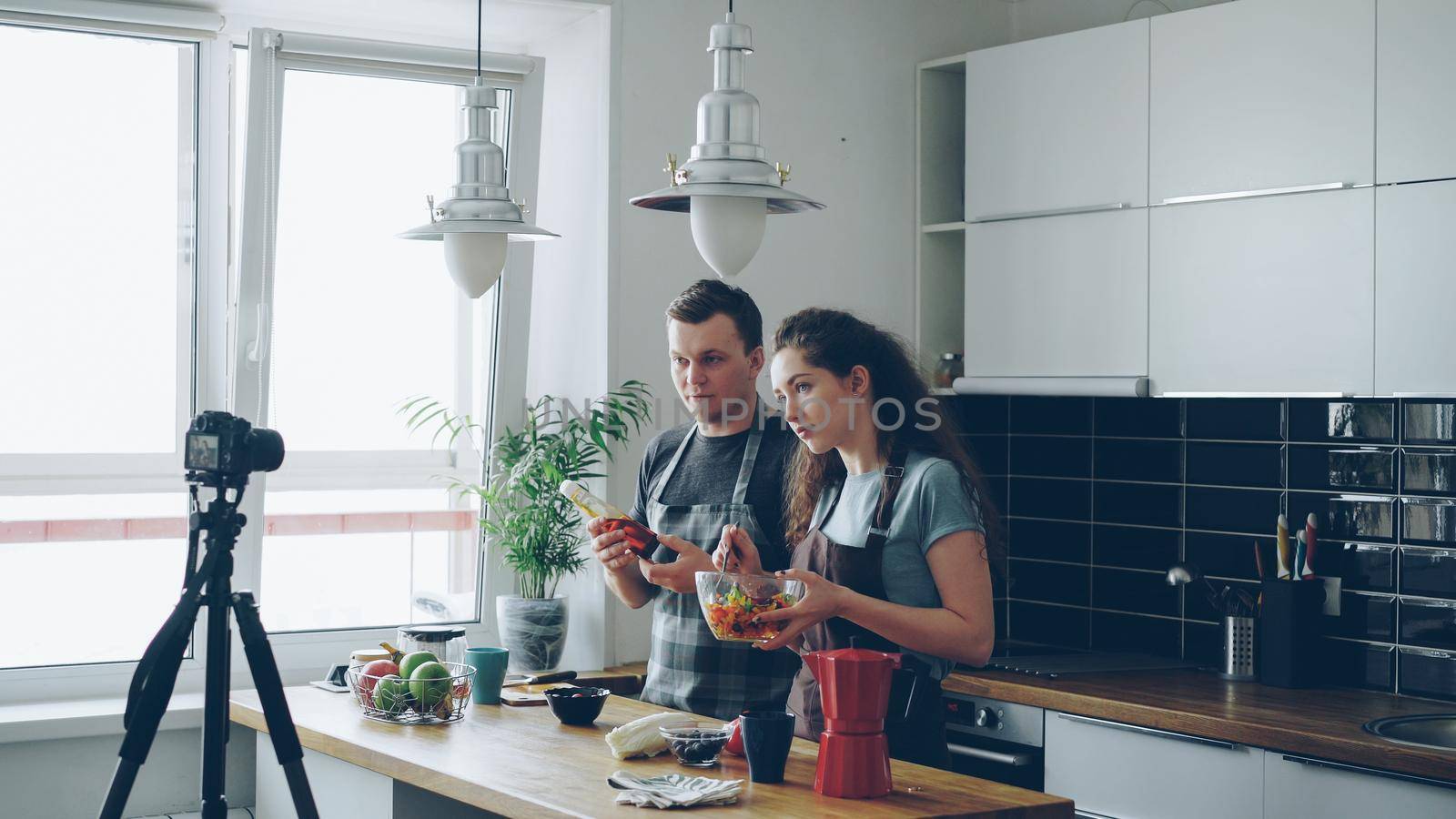 Smiling attractive couple recording video food vlog about cooking haelthy salad on digital camera in the kitchen at home. Vlogging and social media concept