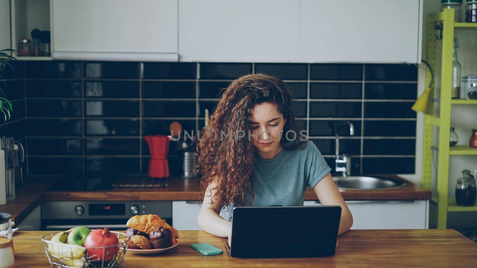 young pretty curly caucasian woman sitting alone at table in nice spacious kitchen idoors working on laptop by silverkblack