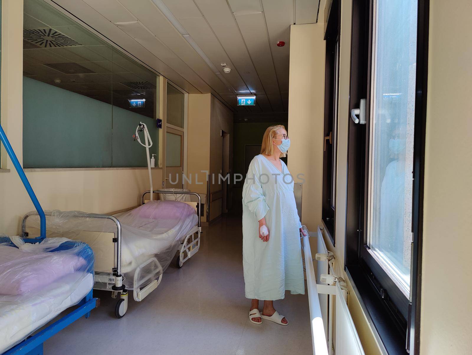 Solitary female patient wearing mask and hospital robe looking out trough big windows of hospital hallway