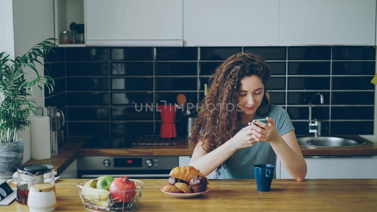 young beautiful curly pretty caucasian woman sitting at table in nice kitchen using smartphone, she is texting someone and smiling, calm and happy by silverkblack