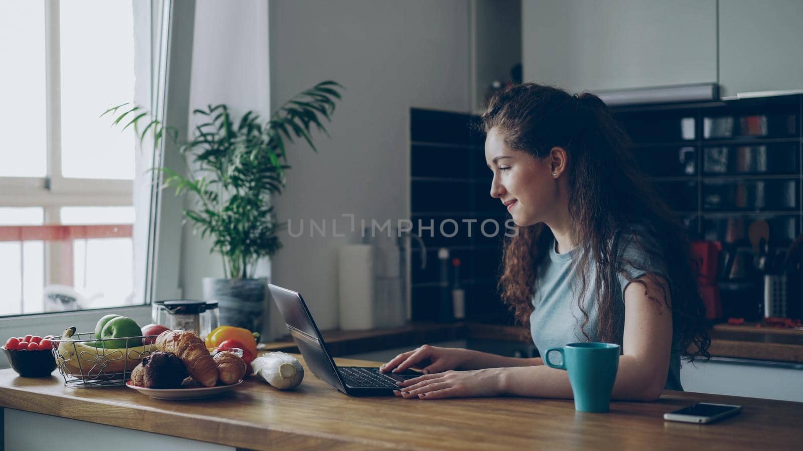 Young concentrated cheerful woman working in kitchen typing laptop computer during breakfast in the morning by silverkblack