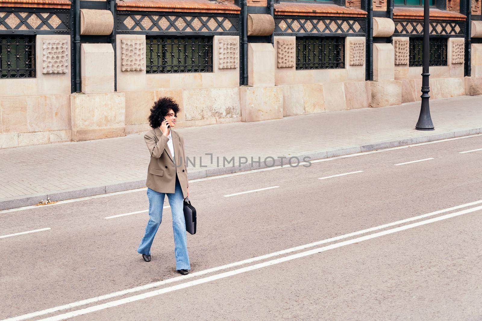 latin business woman walking across the street talking by phone with a briefcase in her hand, concept of urban lifestyle and success, copy space for text