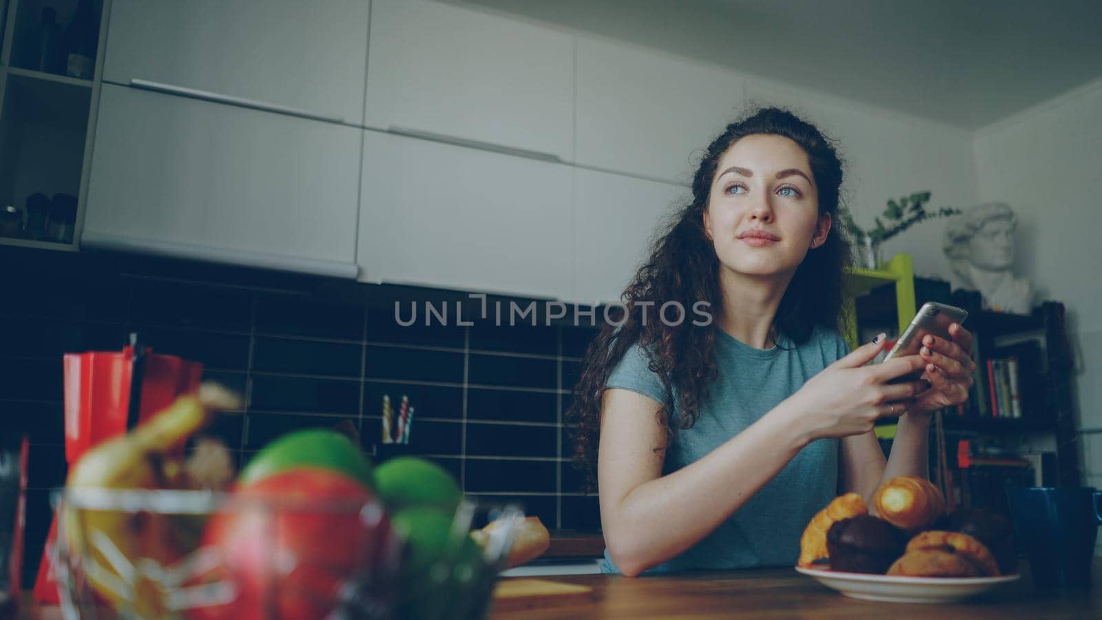 young beautiful curly pretty caucasian woman sitting at table in nice modern kitchen using smartphone, she is texting someone and smiling, calm, happy and positive