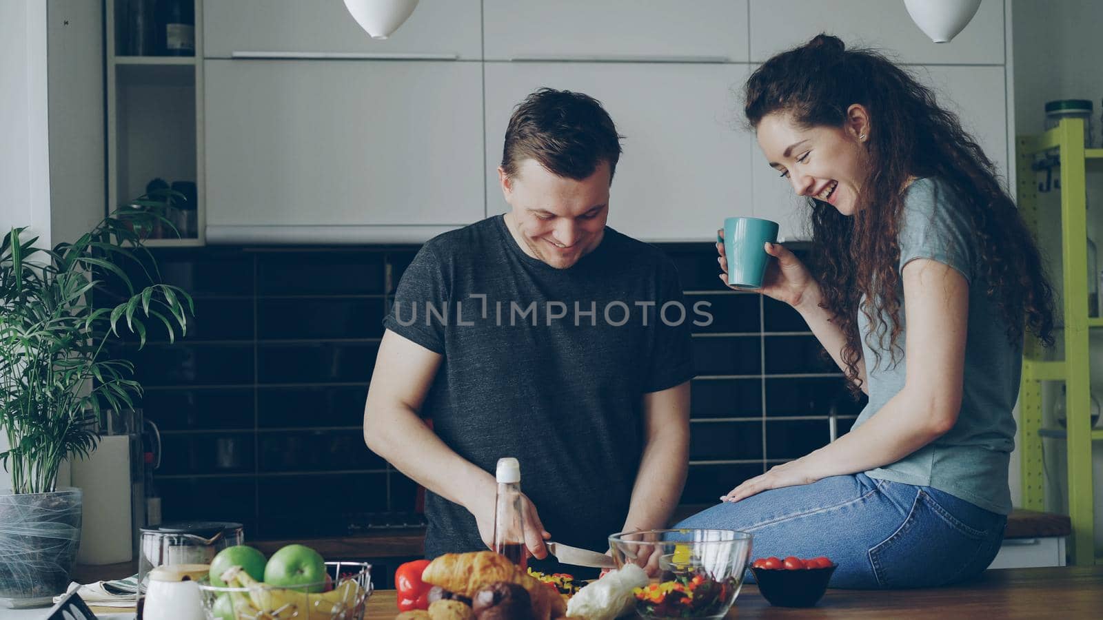 Happy young couple cooking and chatting happily while man cutting vegetables for breakfast in the kitchen at home. Relationship and family concept by silverkblack