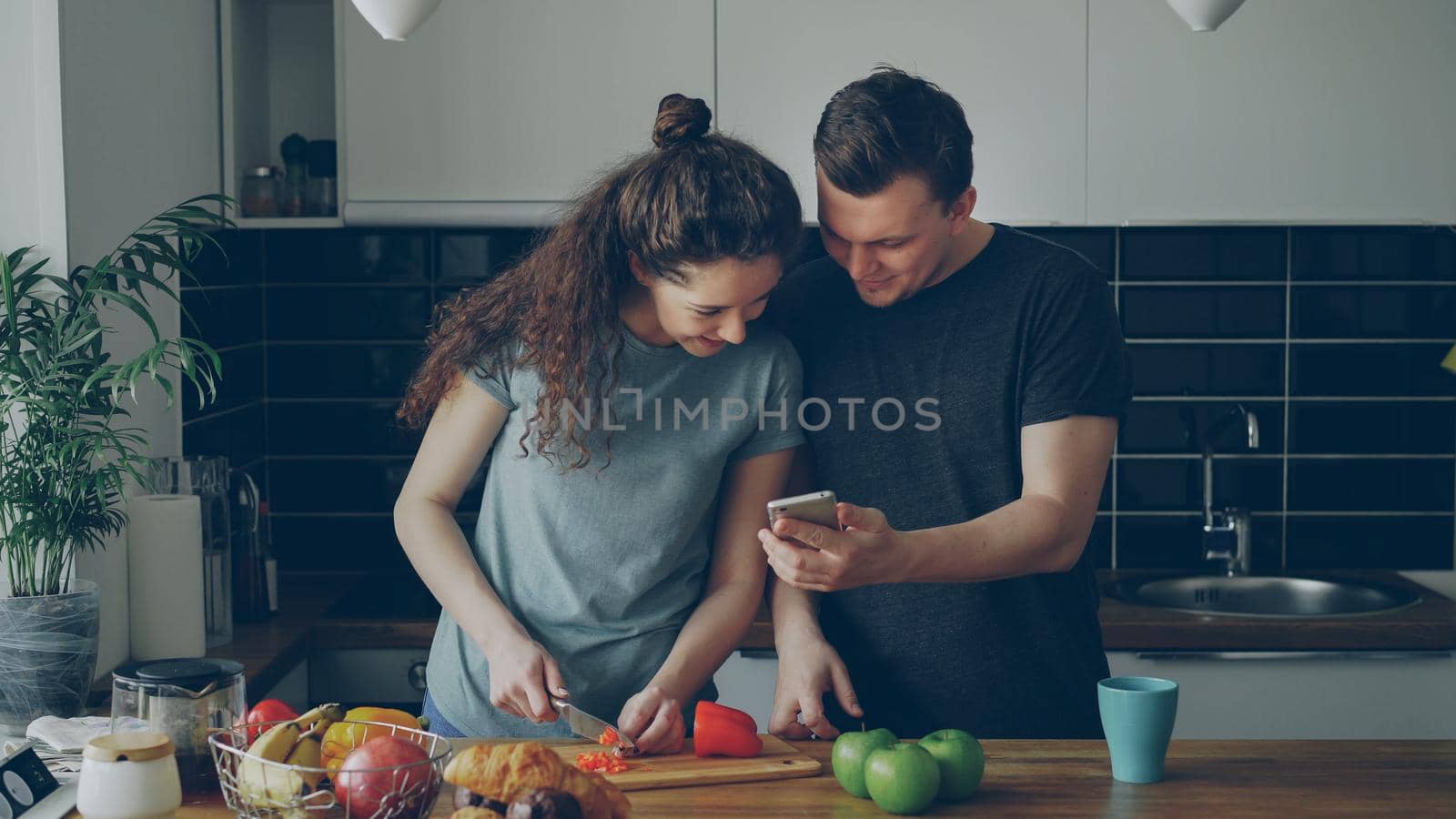 Curly woman near table cutting red pepper in kitchen at home, her boyfriend showing her something funny in smartphone and they are smiling by silverkblack