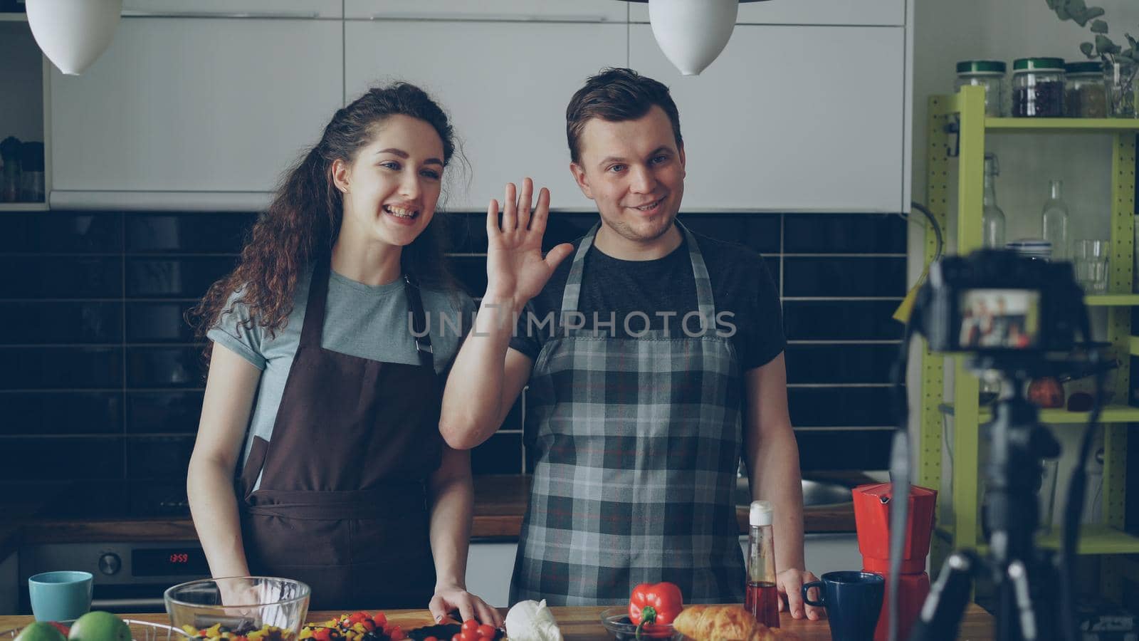 Cheerful attractive couple recording video food vlog about healthy cooking on digital camera in the kitchen at home. Vlogging and social media concept by silverkblack