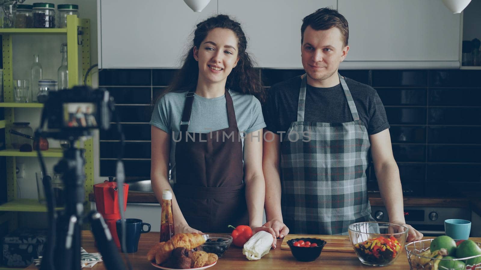 Smiling attractive couple recording video food vlog about cooking haelthy salad on digital camera in the kitchen at home. Vlogging and social media concept