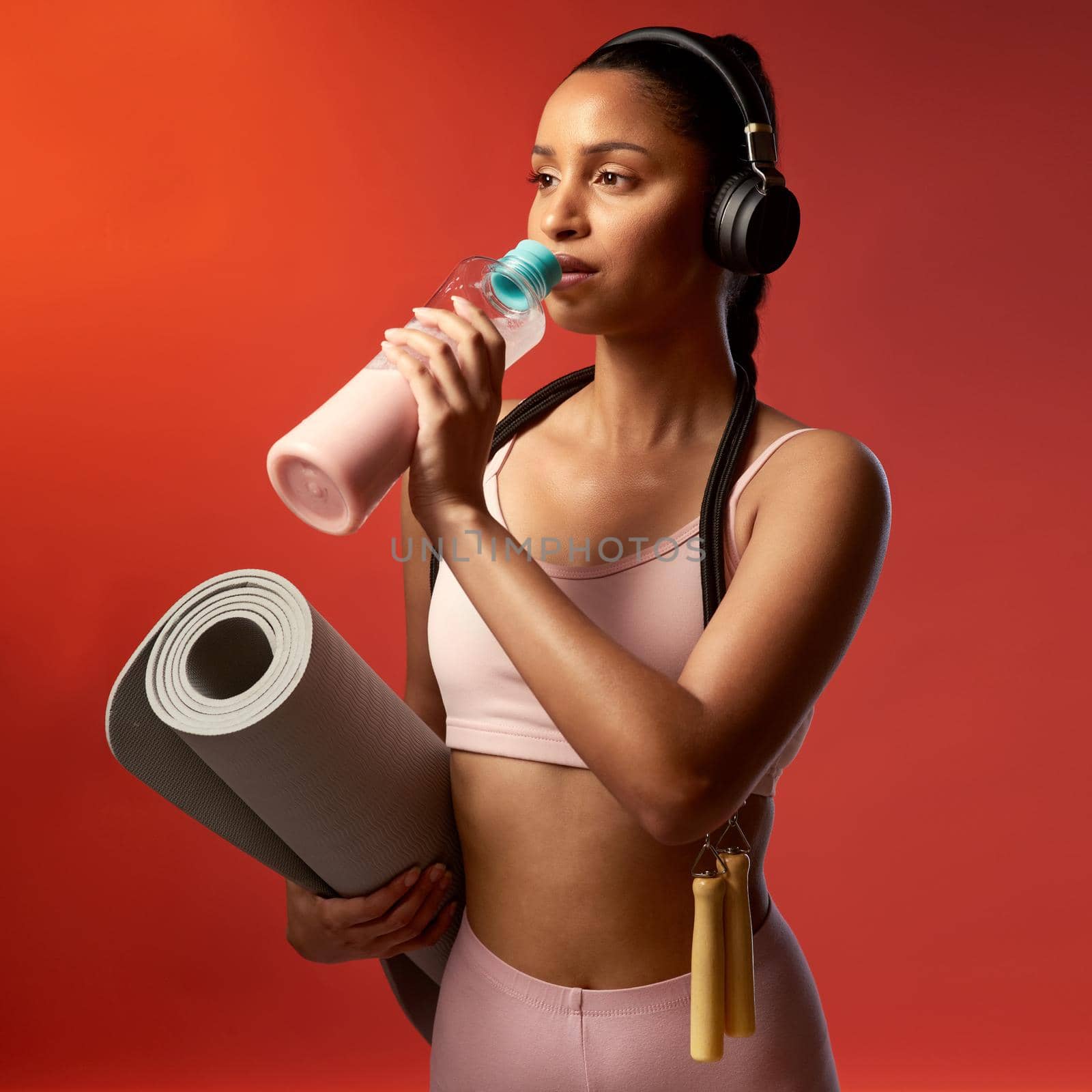 Its not always easy, but its always worth it. Studio shot of a sporty young woman drinking water and holding a yoga mat against a red background. by YuriArcurs