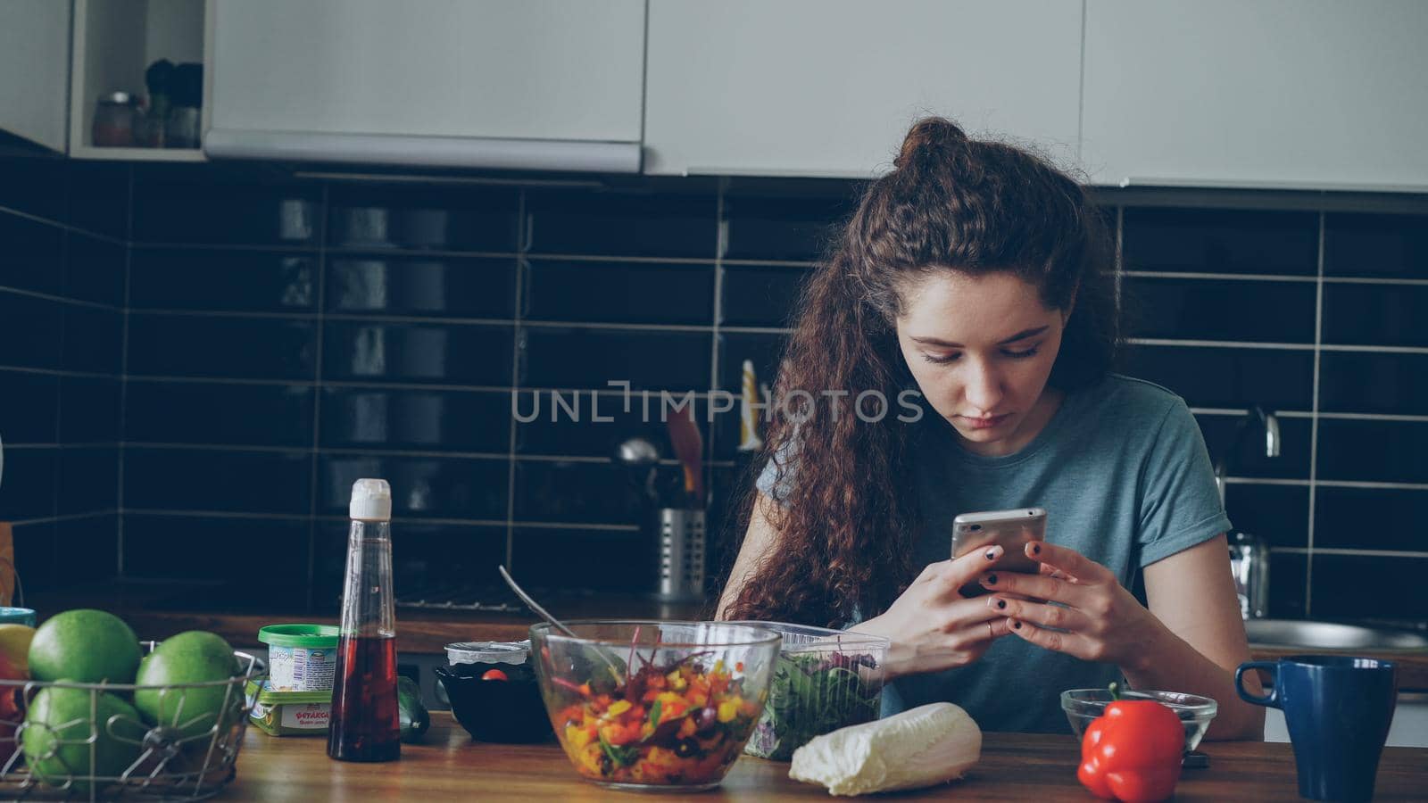 Portrait of young beautiful caucasian woman sitting at table in modern lighty spacious kitchen,using smartphone, texting, plates with food are in front of her