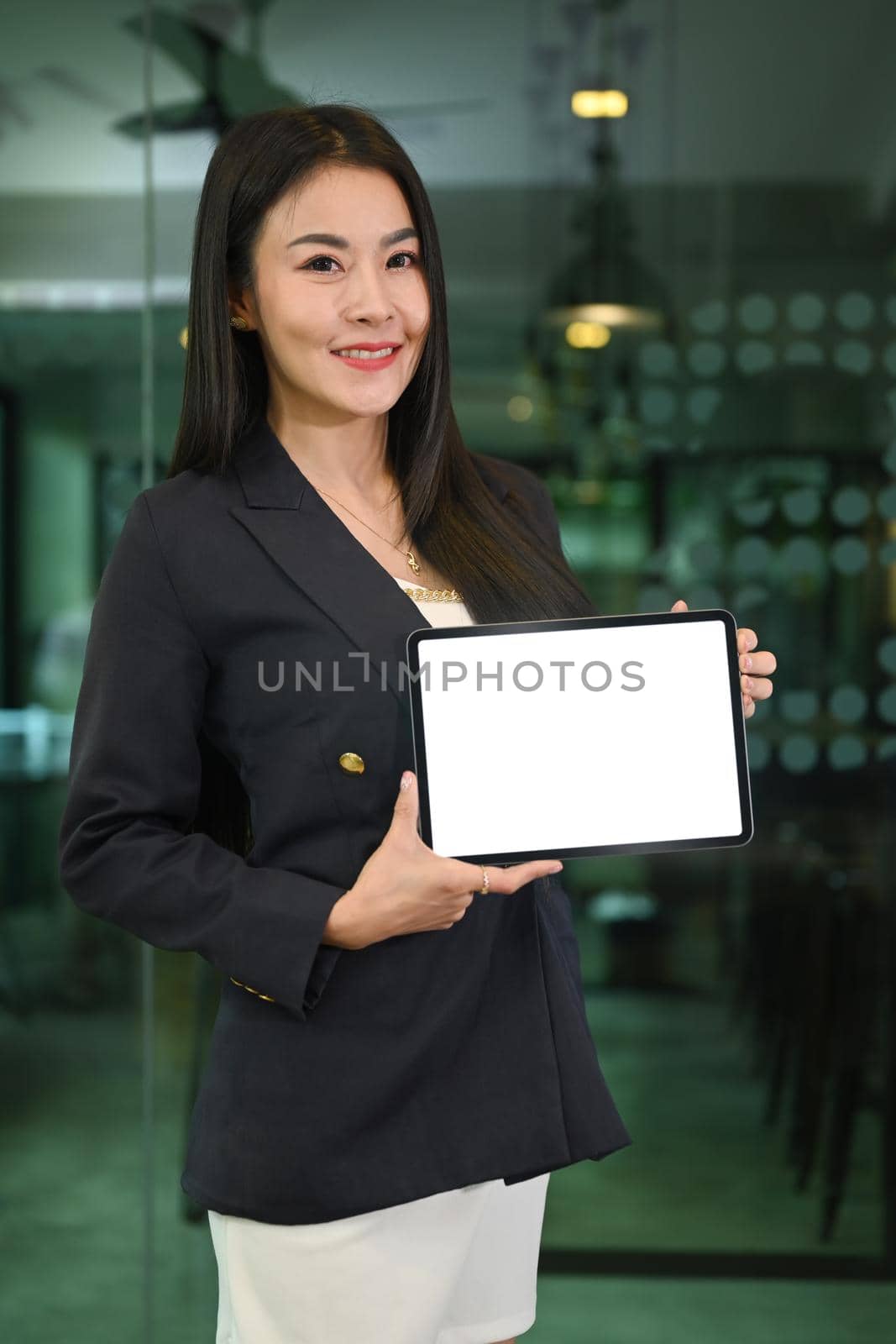 Confident millennial asian woman in business suit holding digital tablet with empty screen and smiling to camera.