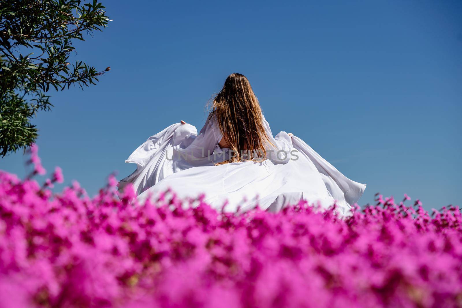 a beautiful woman in a white flowing long dress stay near a beautiful field with pink flowers rear view by Matiunina