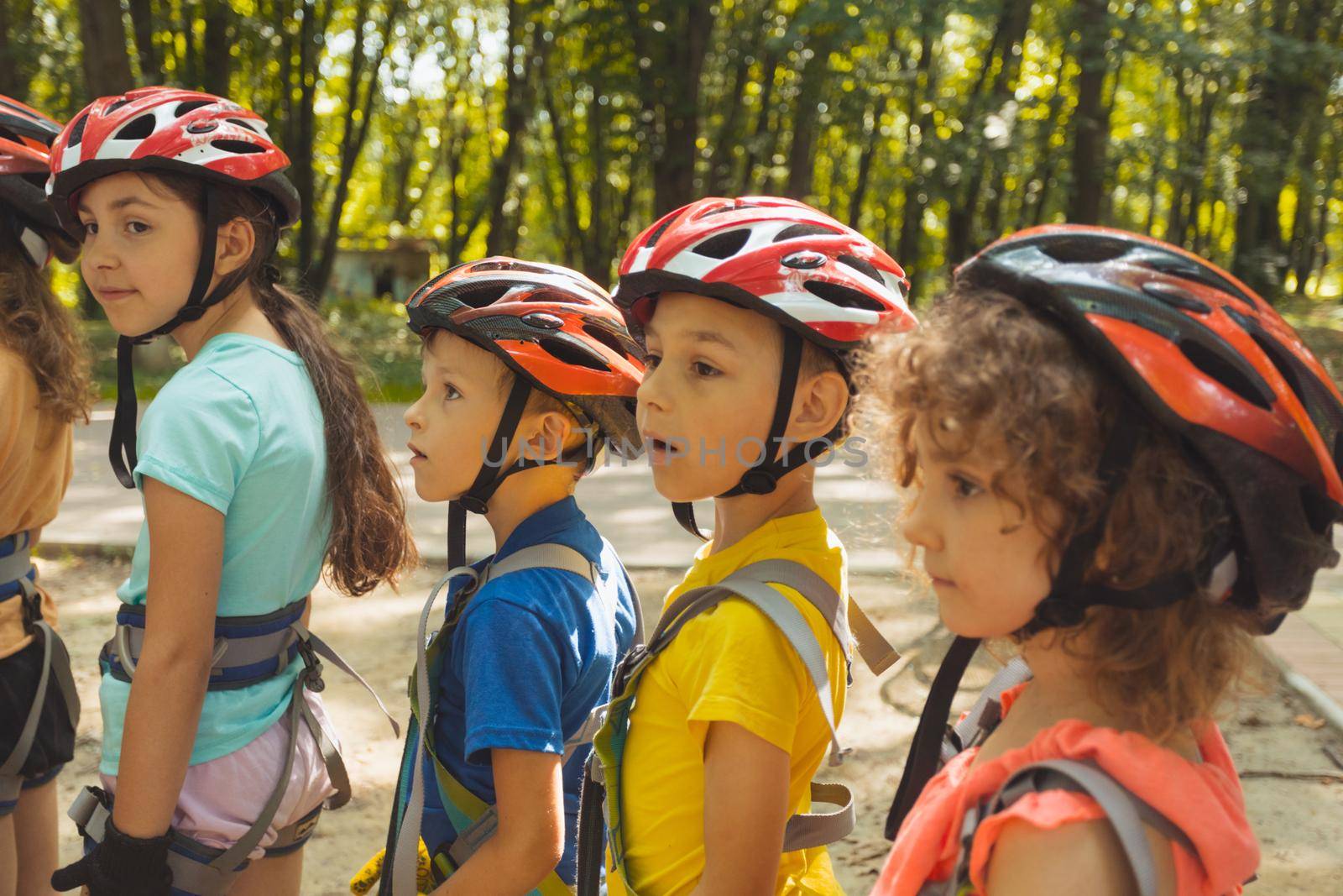 Little boys and girls dressed in protective gear are standing in line for the attraction. The children are standing in line to the rope park and talking