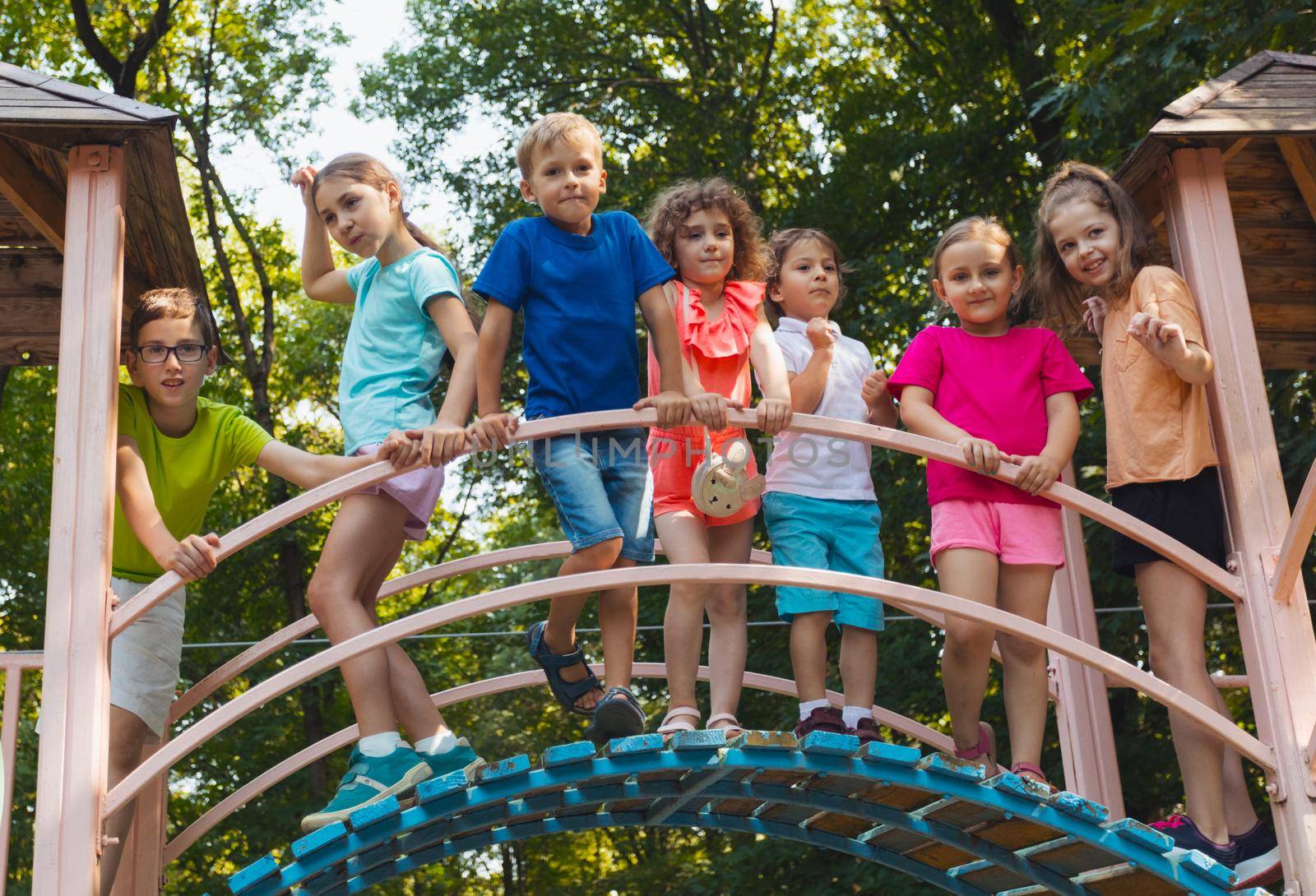 The elementary school children stand on a bridge in the playground and hold on to the railing. The kids dressed in colorful clothes are posing for the camera