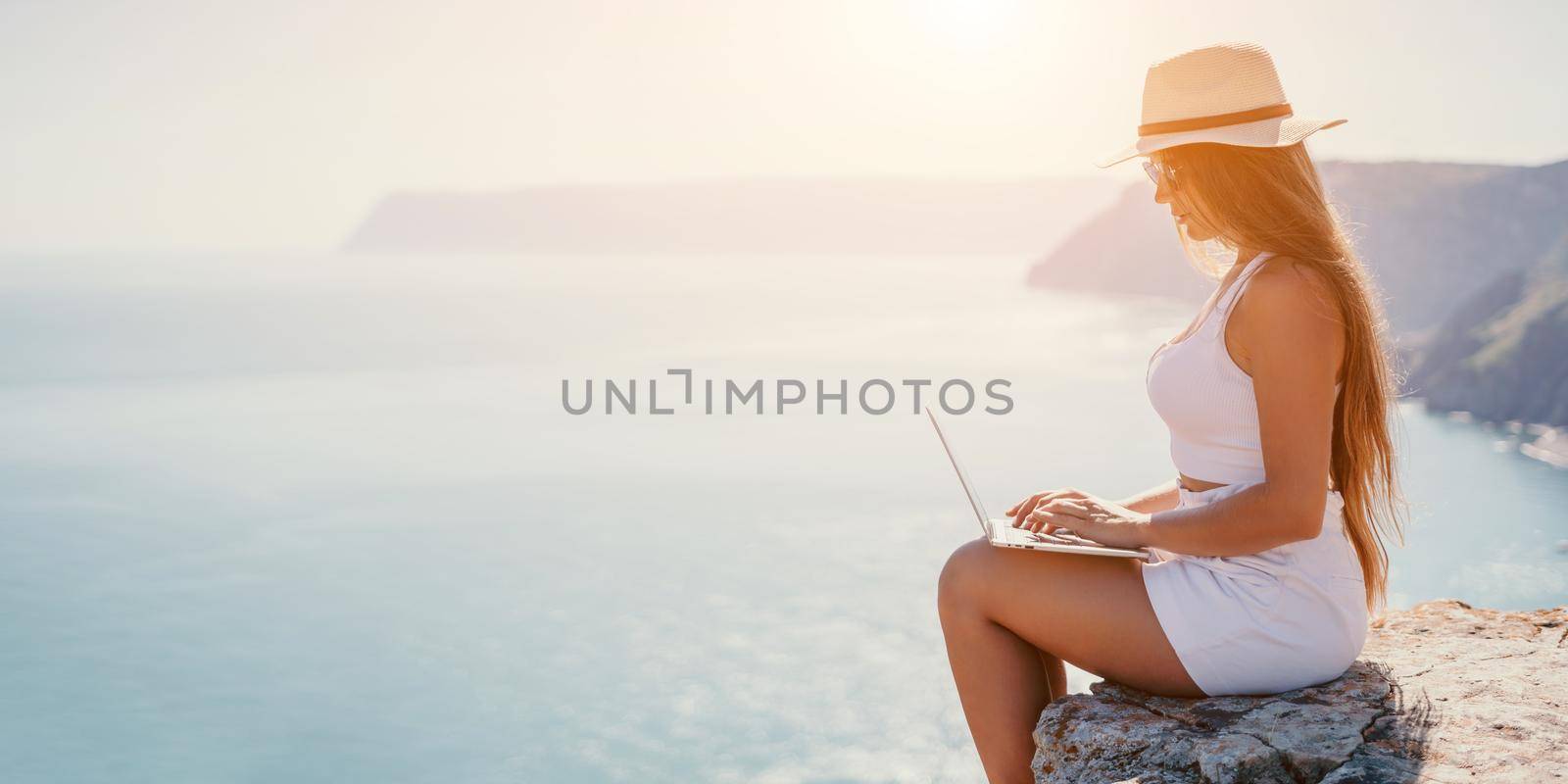 Successful business woman in yellow hat working on laptop by the sea. Pretty lady typing on computer at summer day outdoors. Freelance, travel and holidays concept.