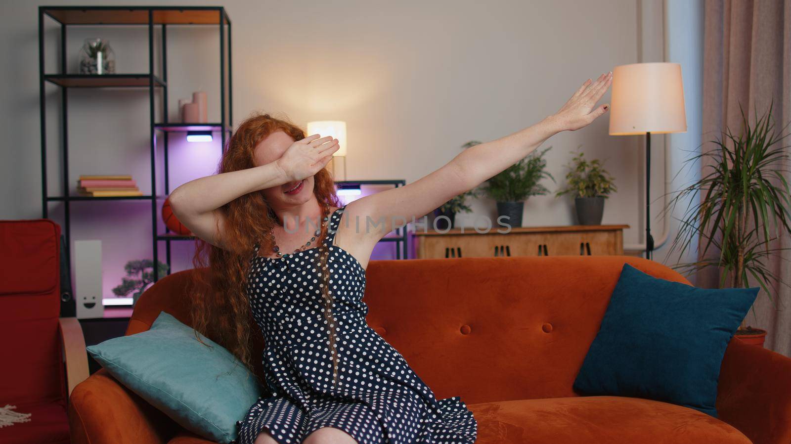 Trendy cheerful positive redhead young woman having fun dancing, moving to rhythm, dabbing raising hands, making dub dance gesture at modern home apartment. Ginger pretty girl in living room on sofa