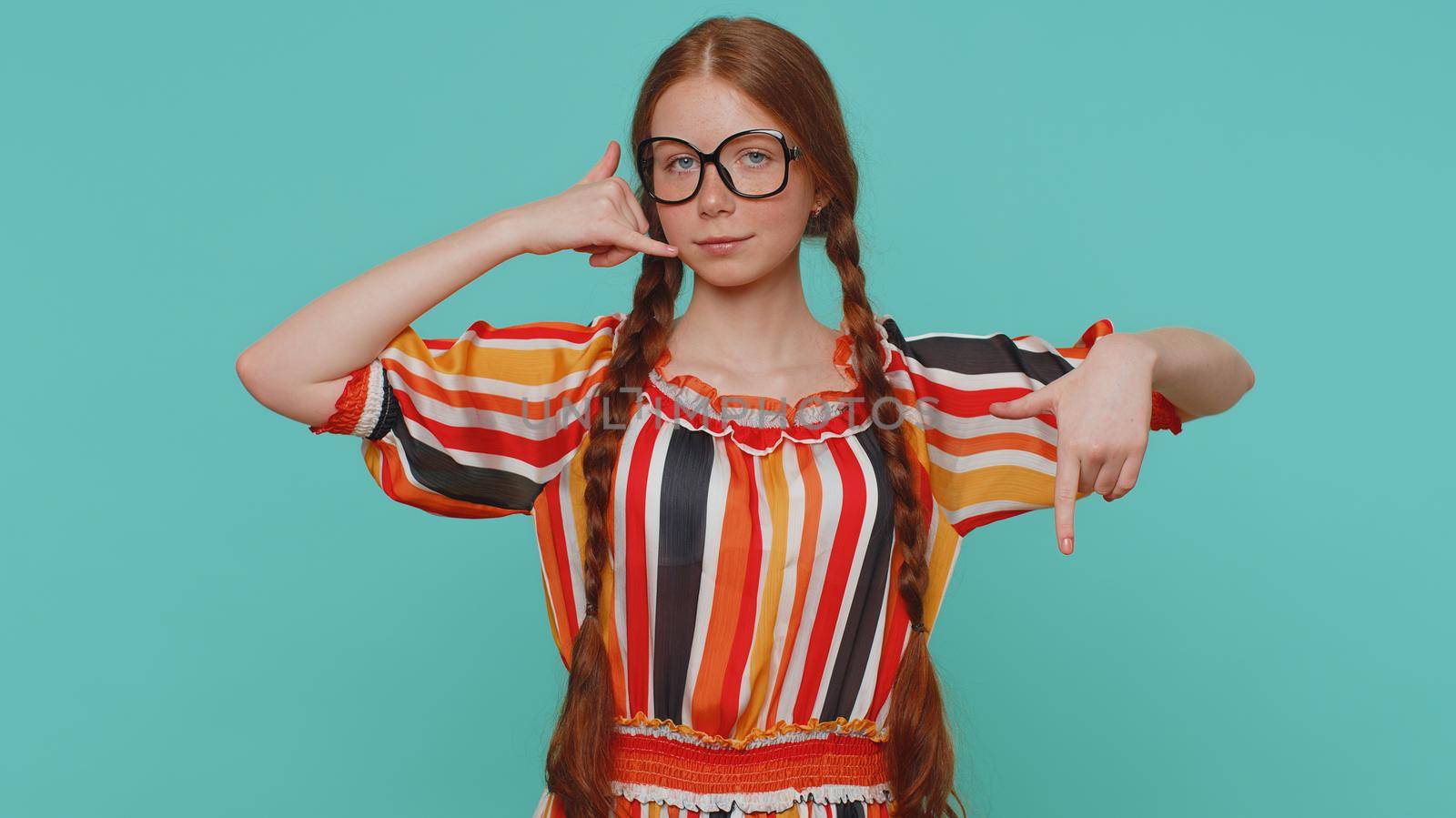 Call me, here is contact number. Redhead girl in dress and glasses looking at camera doing phone gesture like says hey you call me back. Young teenager ginger child kid isolated on blue background