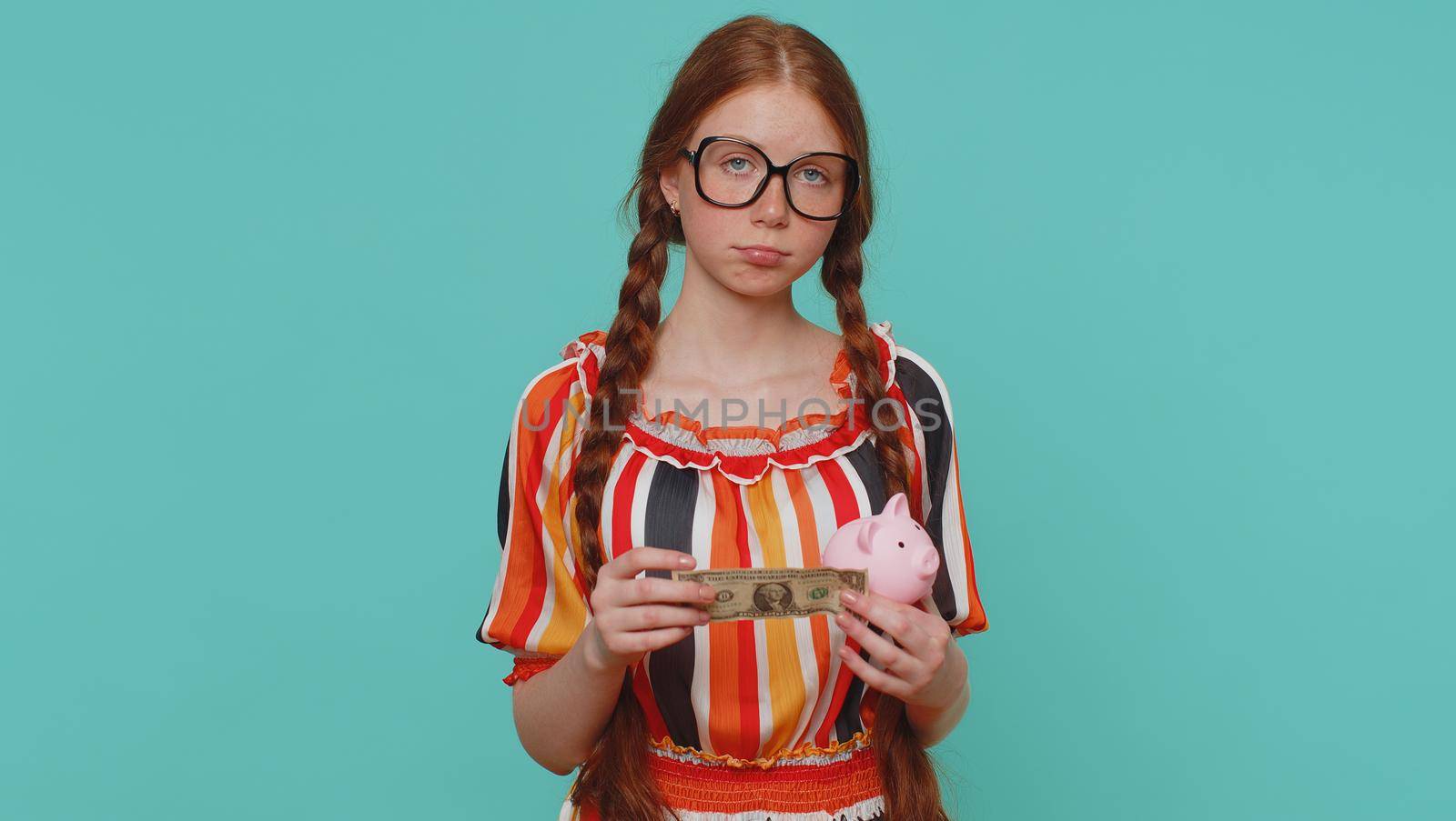 Poor girl upset over insufficient amount of money, holding piggybank and one dollar banknote. Financial crisis. Bankruptcy. Poverty and destitution. Young teenager child on blue studio background