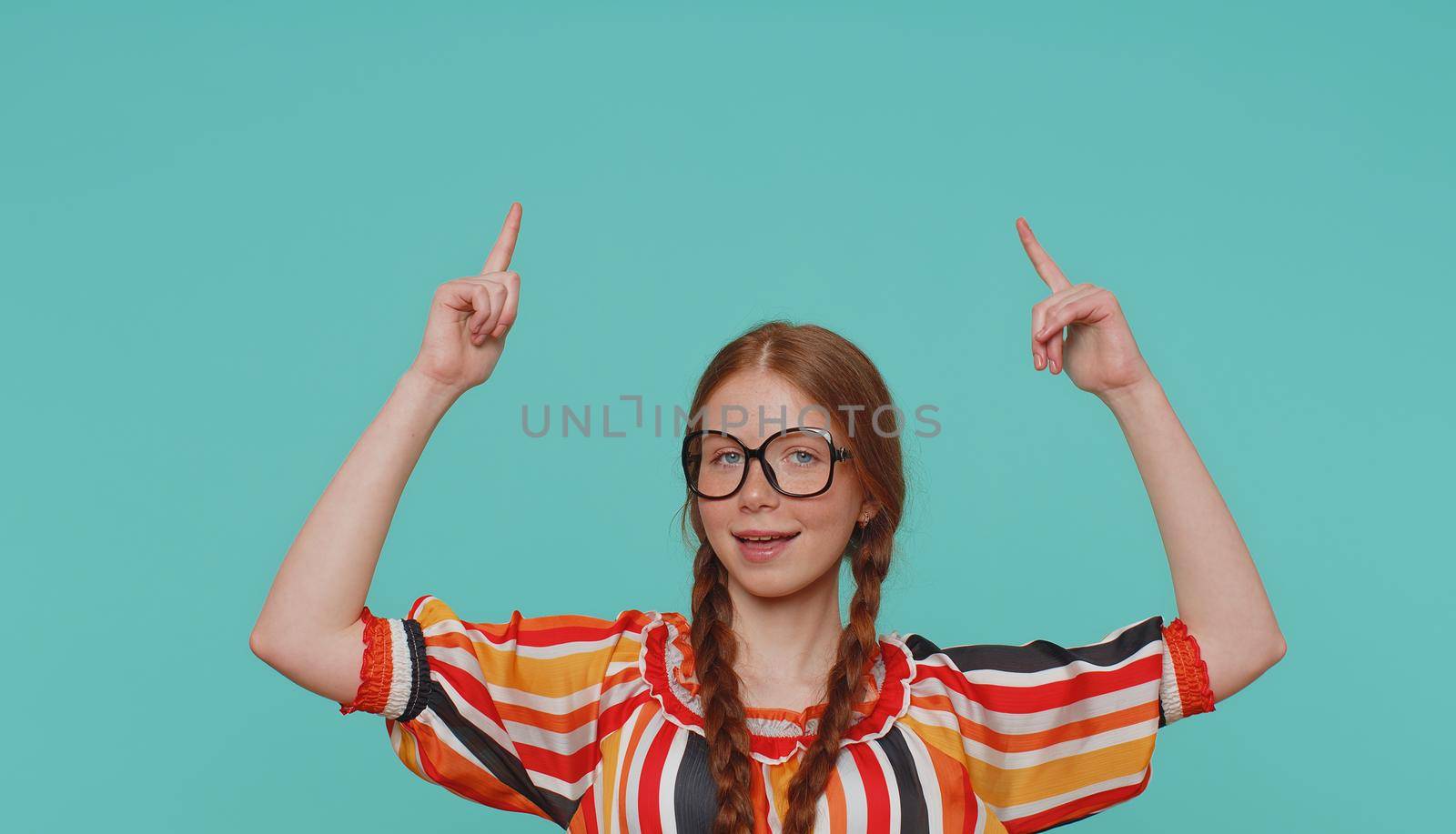 Beautiful redhead girl kid showing thumbs up and pointing up empty place, advertising area for commercial text, copy space for goods promotion. Young ginger teenager child on blue studio background