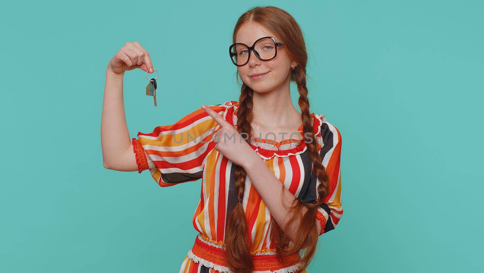 Redhead young girl real estate agent lifting hand showing the keys of new home house apartment, buying or renting property, mortgage loan. Red hair one ginger teenager child on blue studio background
