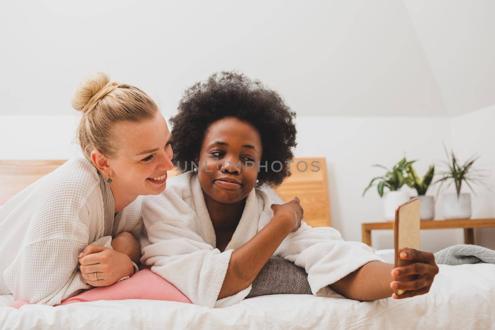Two young women are lying on the bed and take selfies. The women in bathrobes are resting after spa treatments