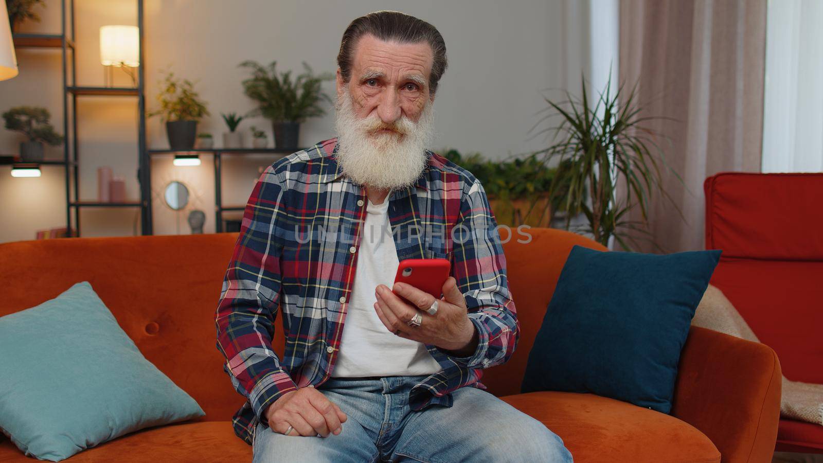 Senior old grandfather sitting on sofa uses mobile phone smile at modern home apartment. Elderly man texting share messages content on smartphone social media applications online, watching relax movie