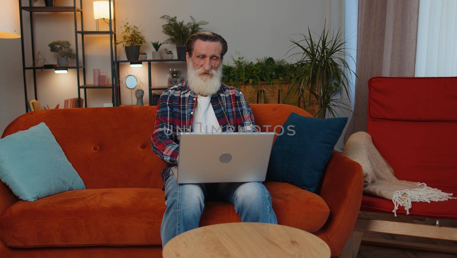 Senior old grandfather man freelancer at home room couch working on laptop. Elderly retired grandpa works on notebook, sends messages, makes online order purchases or food delivery, watching movies