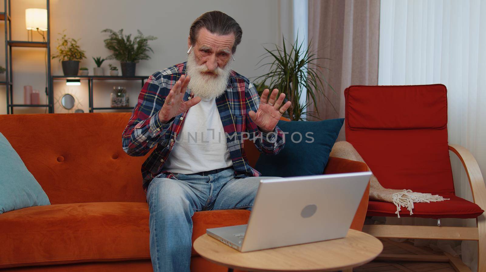Senior old grandfather sitting on couch, looking at laptop, making video webcam conference call with friends or family, enjoying pleasant conversation. Elderly man laughing, waving hello alone in home
