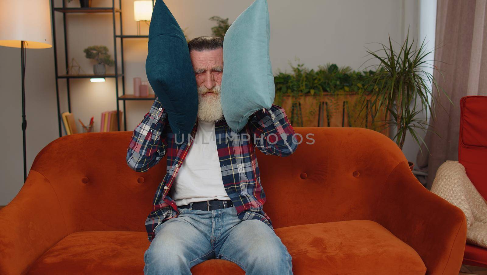 Repair work at neighbours. Irritated senior man sit on couch cover ears with pillows annoyed by noisy neighbors suffer from headache wishes silence. Thin walls at home flat without sound insulation
