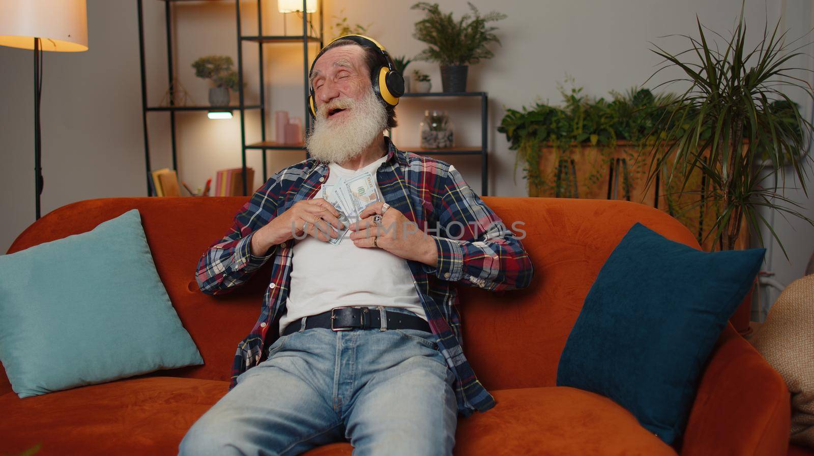 Successful rich senior businessman grandfather man holding money cash cheering up with high profits, lottery game win, satisfied of wealth income earnings salary on home couch, dancing listening music