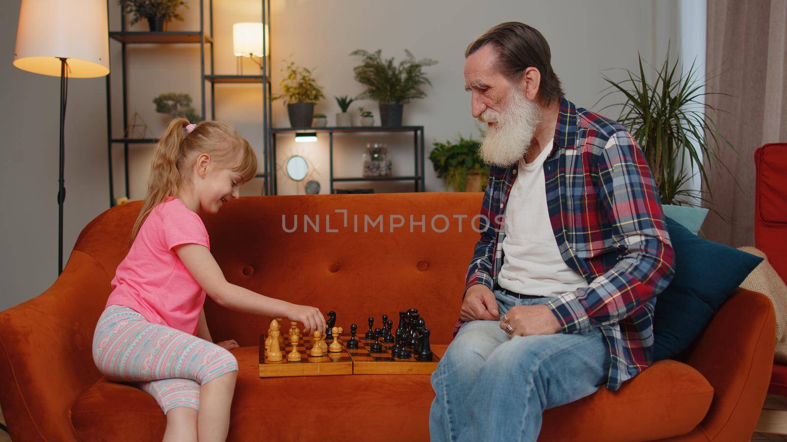 Smiling senior old grandfather man playing chess with teen granddaughter at home living room. Retired elderly grandparent enjoying play board game, resting with child girl kid. Family leisure activity