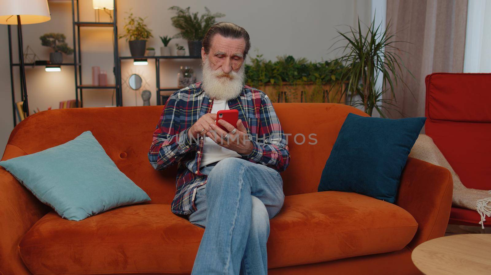 Senior old grandfather sitting on sofa uses mobile phone smile at modern home apartment. Elderly man texting share messages content on smartphone social media applications online, watching relax movie