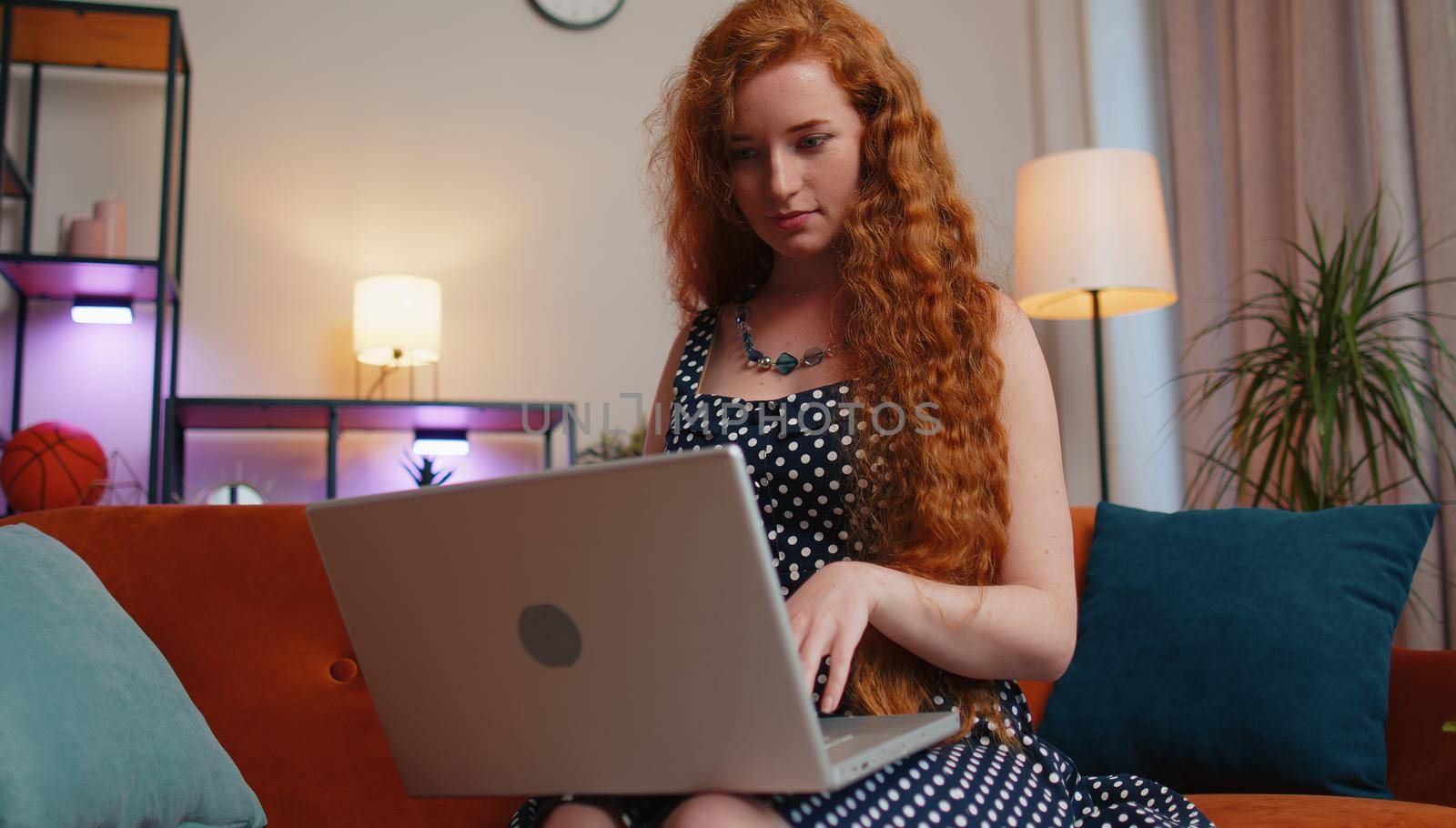Redhead young woman in polka dots dress freelancer in home living room sits on couch, opens laptop start working. Ginger girl works on notebook, sends messages, makes online purchases, watching movies