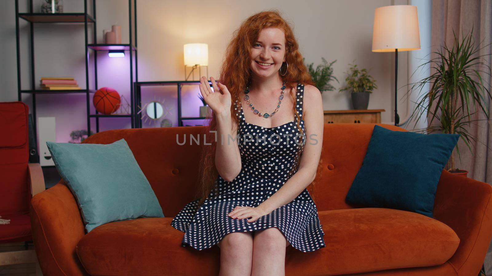 Beautiful adult girl smiling friendly at camera and waving hands gesturing hello, hi, greeting or goodbye, welcoming with hospitable expression. Young redhead woman indoors at home in room on couch