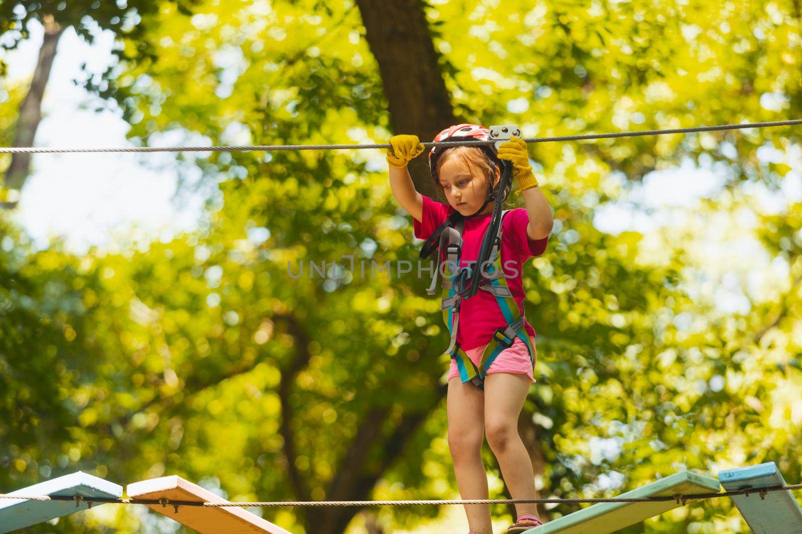 The concentrated little girl carefully steps on the suspension bridge. The girl in protective gear is playing in the rope park