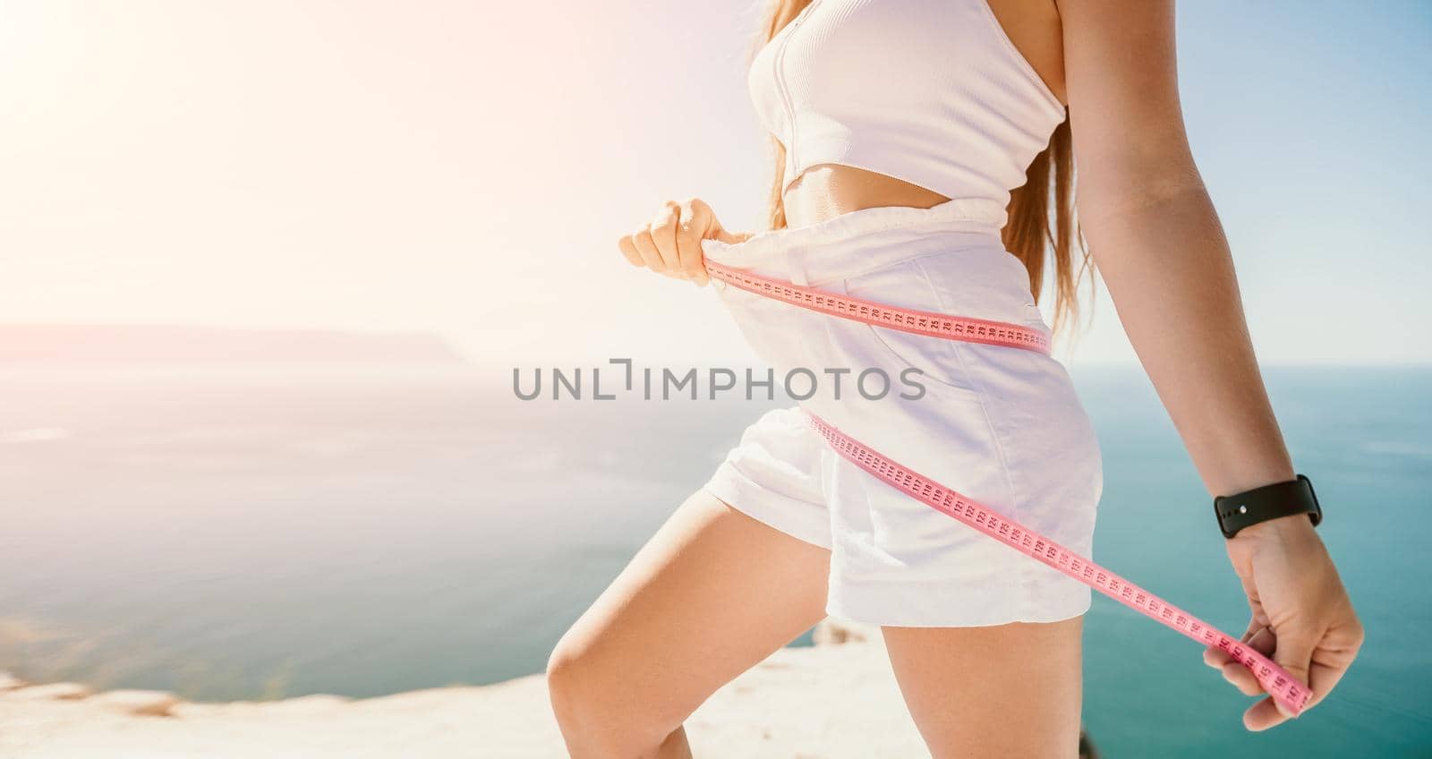 woman in white shorts holds in her hands pink measuring tape on her waist near the azure sea. Concept of detox and diet by panophotograph