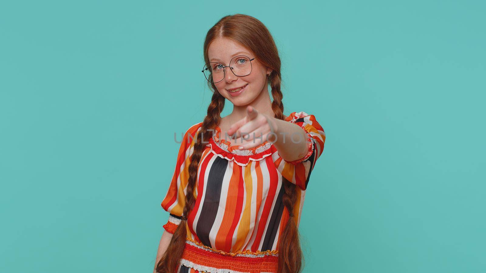 I choose you. Redhead girl pointing to camera and looking with playful happy expression, making choice, showing direction. Young one teenager ginger child kid indoor isolated on blue studio background
