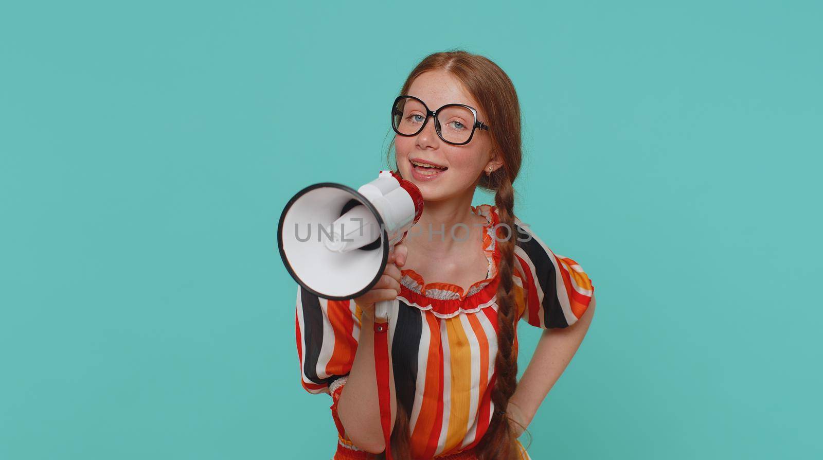 Smiling happy redhead girl talking with megaphone, proclaiming news, loudly announcing sale advertisement, warning using loudspeaker to shout speech. Young red hair teenager child on blue background