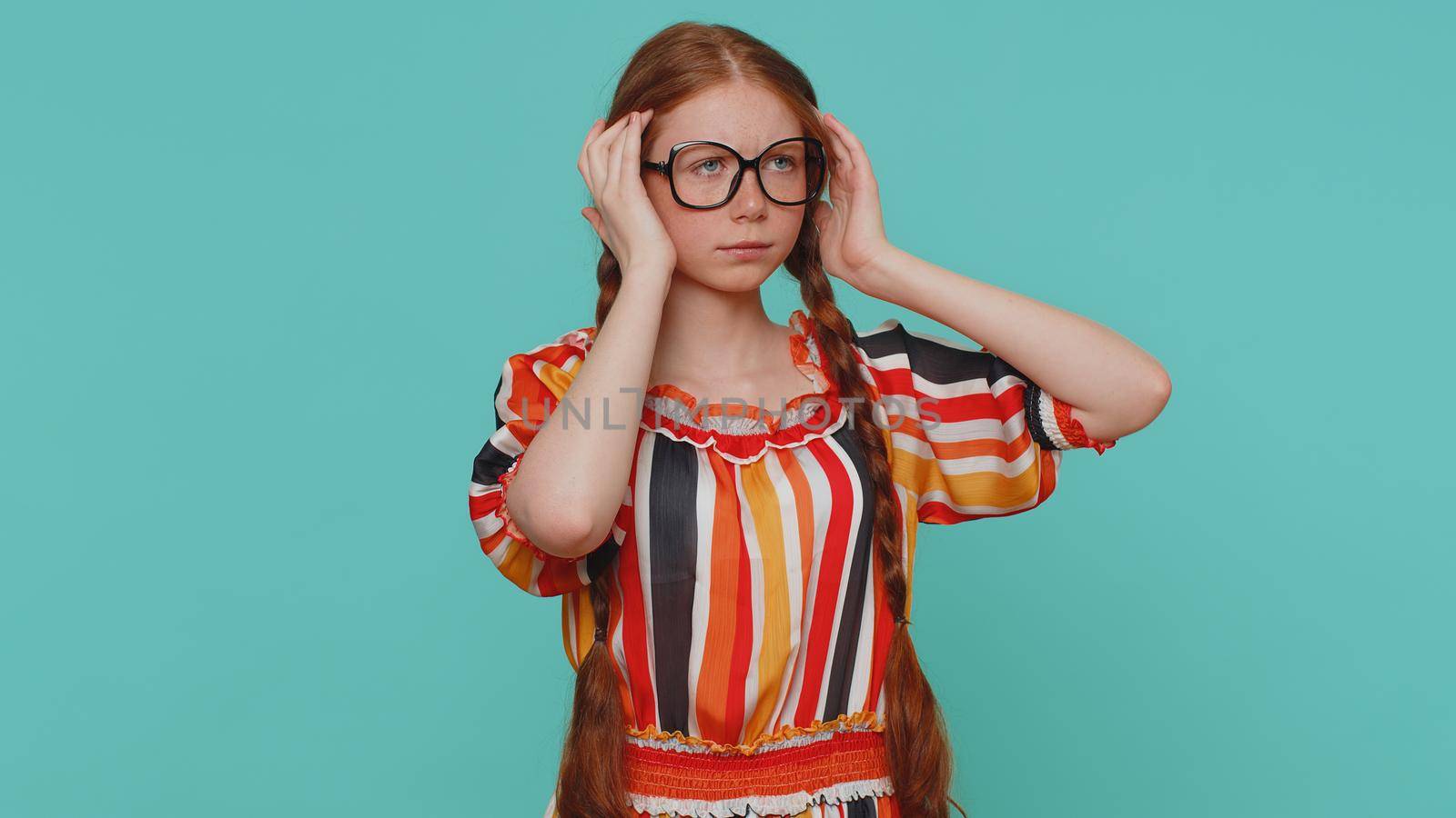 Redhead young girl in dress rubbing temples to cure headache problem, suffering from tension and migaine, stress, grimacing in pain, high blood pressure. Child isolated alone on blue studio background