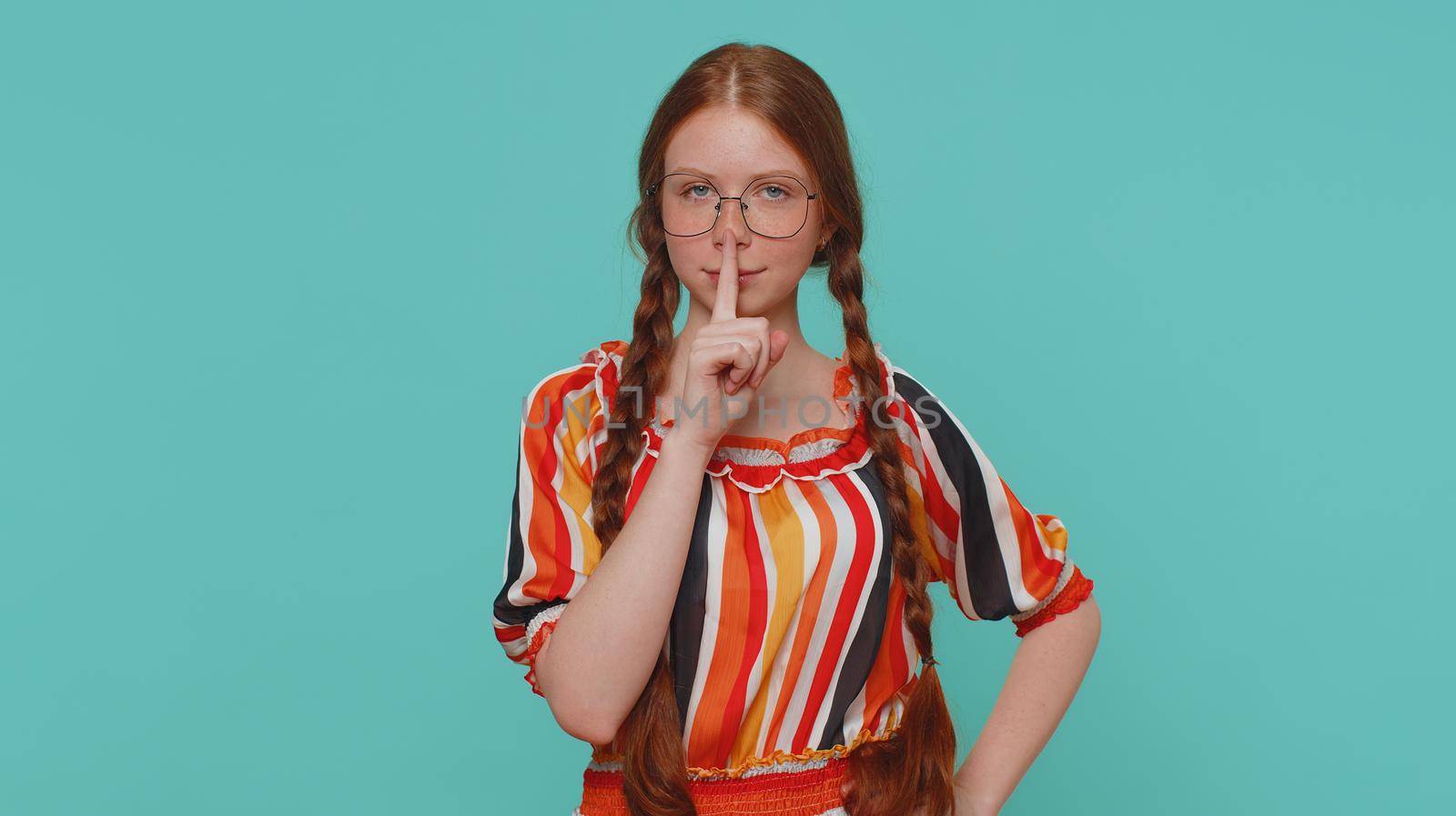 Shh be quiet please. Portrait of redhead girl 20s presses index finger to lips makes silence gesture sign do not tells secret. Young beautiful ginger teenager child isolated on blue studio background