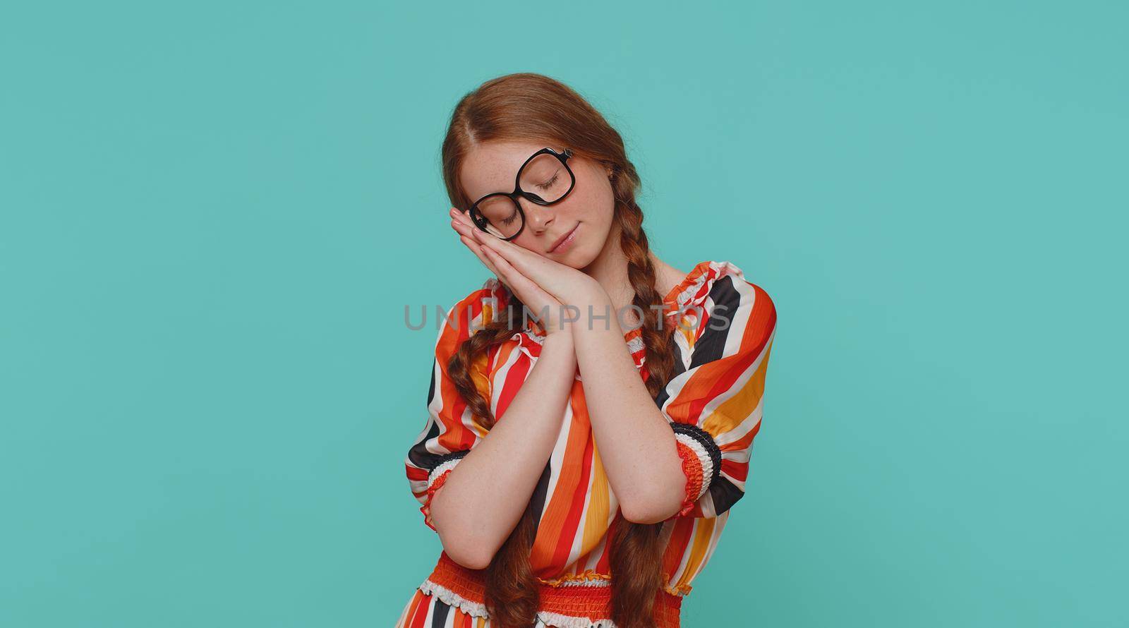 Tired redhead girl going to sleep laying down on his palms, yawning, dozing off during day, taking nap and resting, comfortable bedtime. Teenager ginger child kid isolated on blue studio background