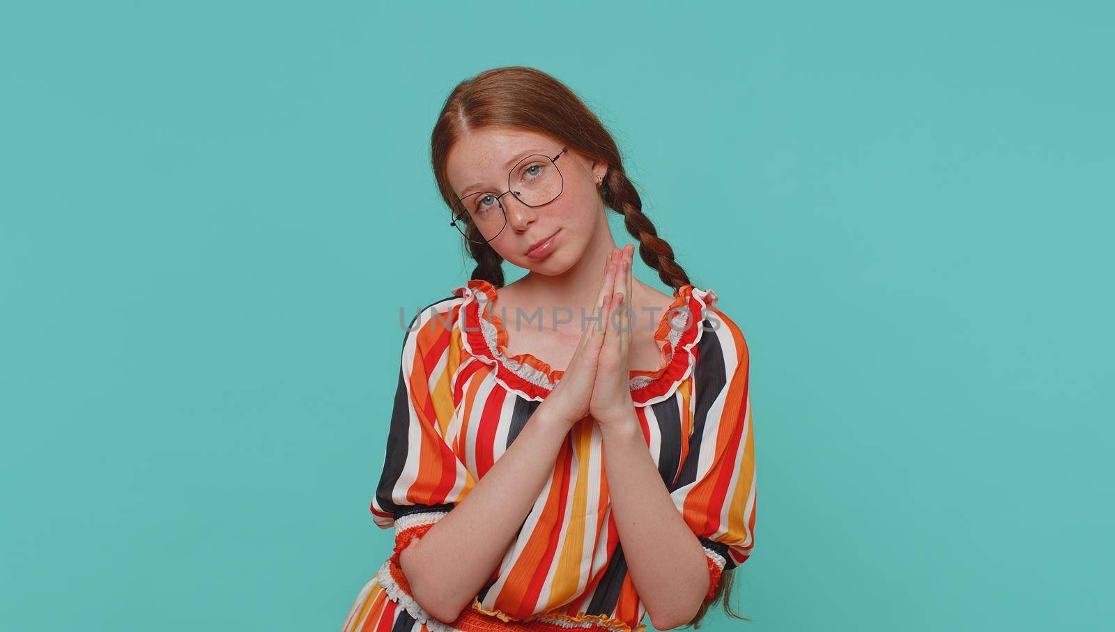 Please, God help me. Redhead girl praying, looking upward and making wish, asking with hopeful imploring expression, begging apology. Young teenager ginger child kid isolated on blue studio background