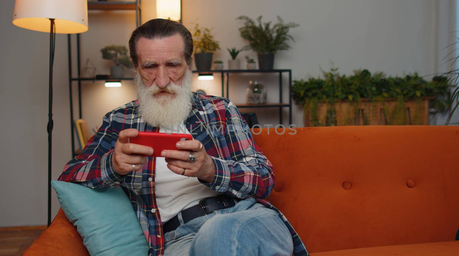Worried senior old grandfather man enthusiastically playing racing shooter online video games on mobile phone. Elderly grandpa using smartphone gadget app with drive simulator at home sitting on couch